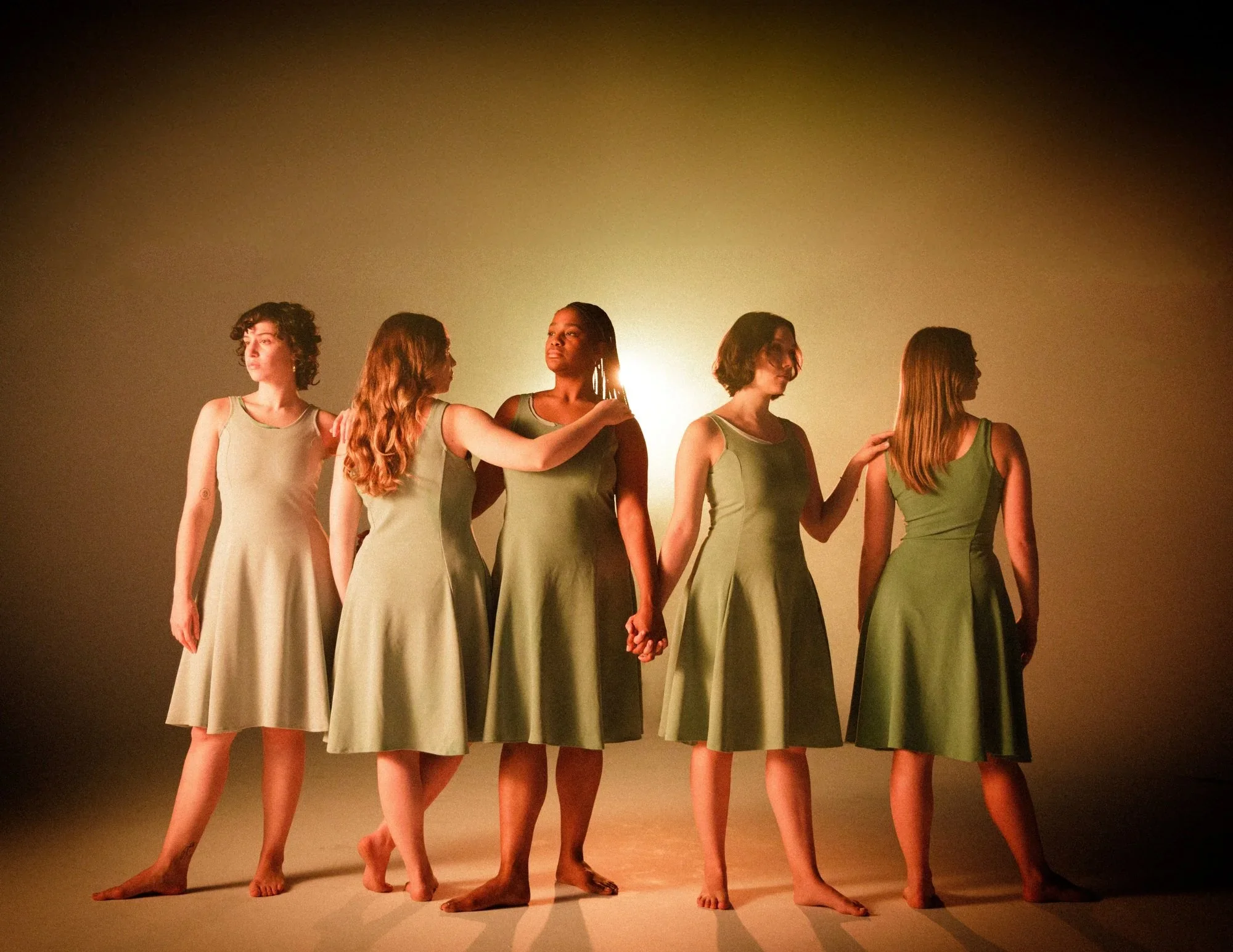 Five women in matching knee-length green dresses standing in a line with their arms around each other's shoulders and holding hands, illuminated by a warm backlight in a minimalist setting.