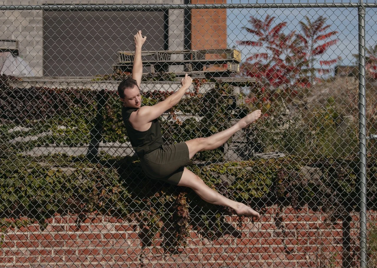 A woman in a black tank top and olive green shorts appears to be hanging or climbing on a chain-link fence, with her arms and legs extended in different directions, in an outdoor urban setting with brick and plants.