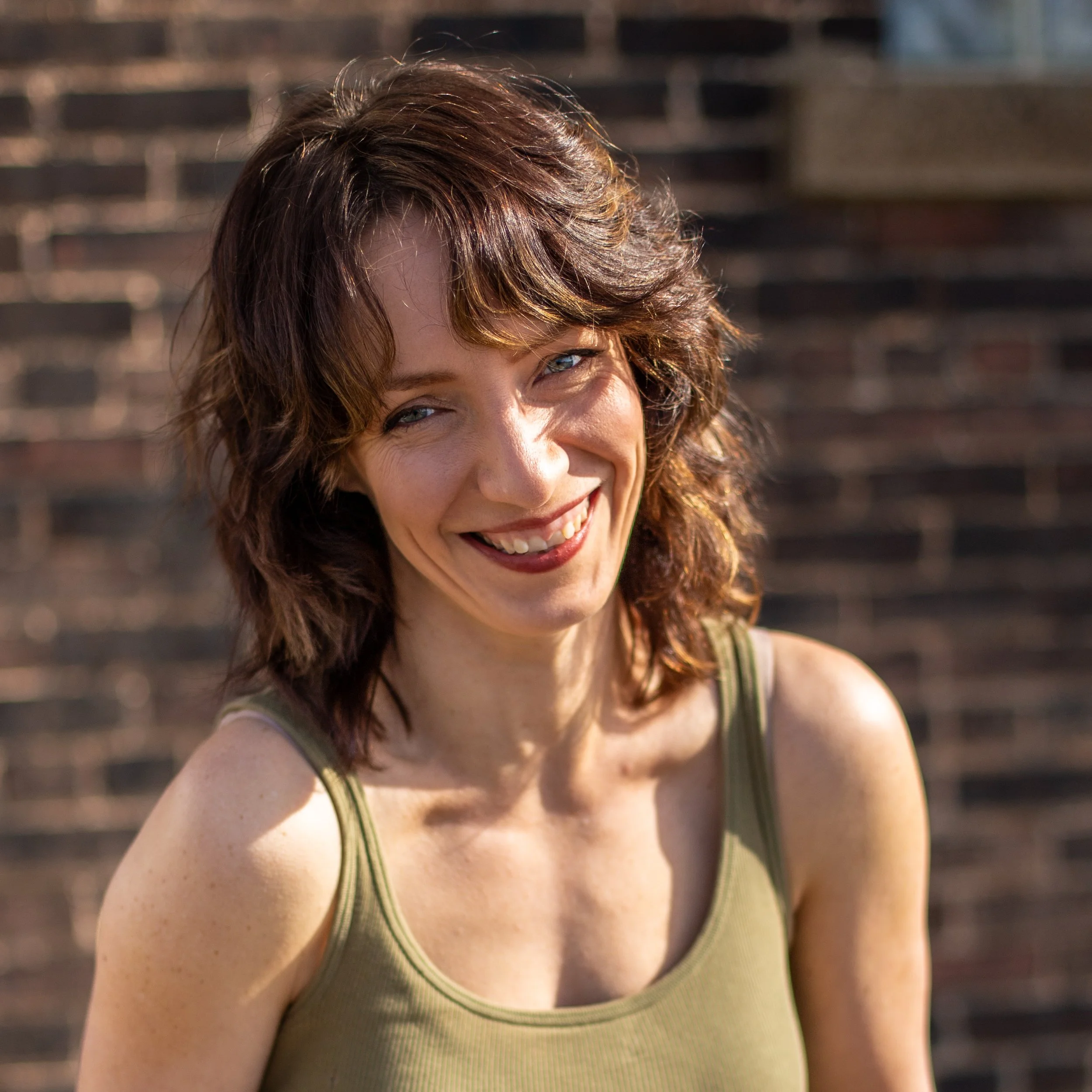 Smiling woman with short, wavy brown hair and blue eyes, wearing a green tank top, standing outdoors in front of a brick wall.