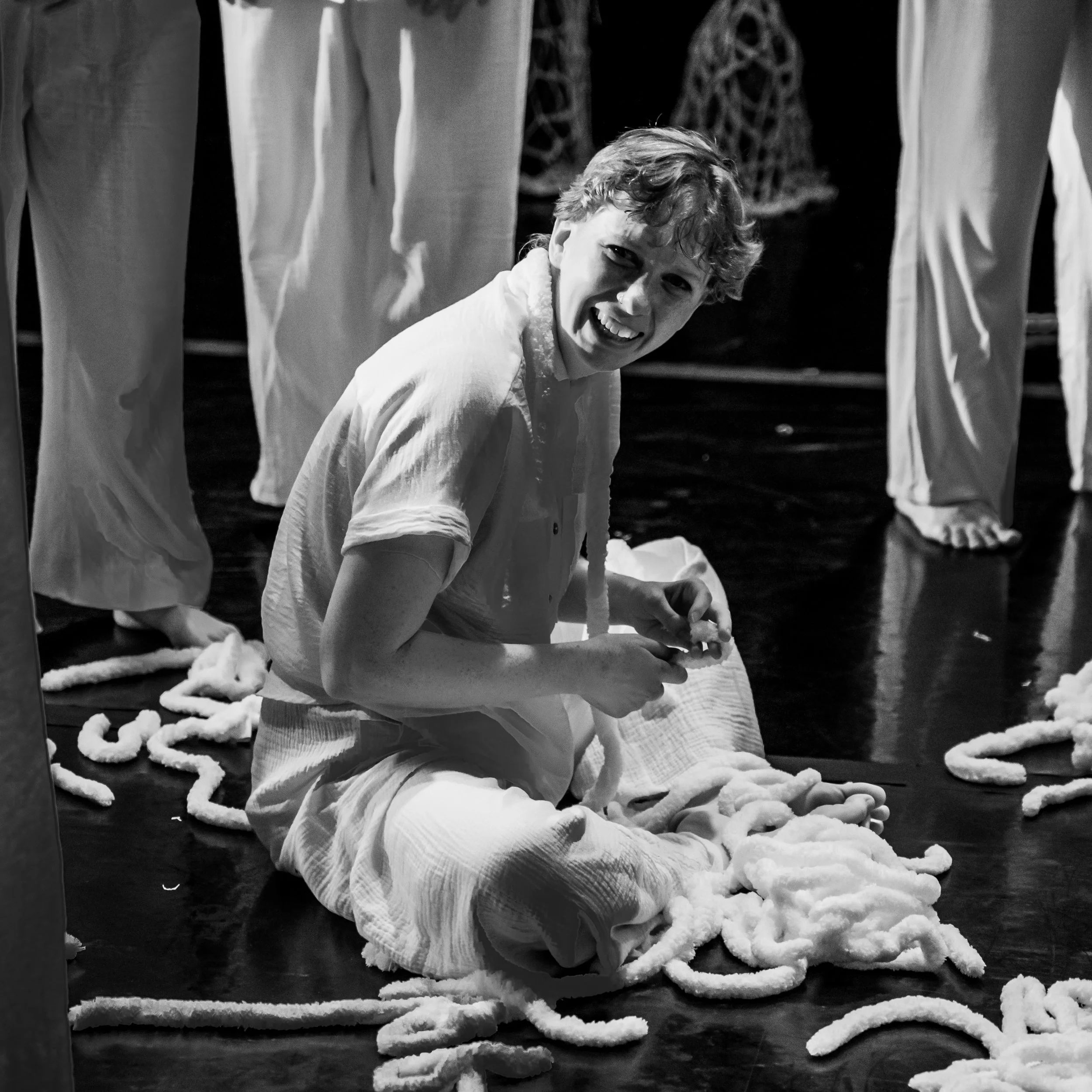 A person sitting on the floor surrounded by twisted lengths of fabric or ropes, smiling or laughing, in a theatrical or artistic setting.