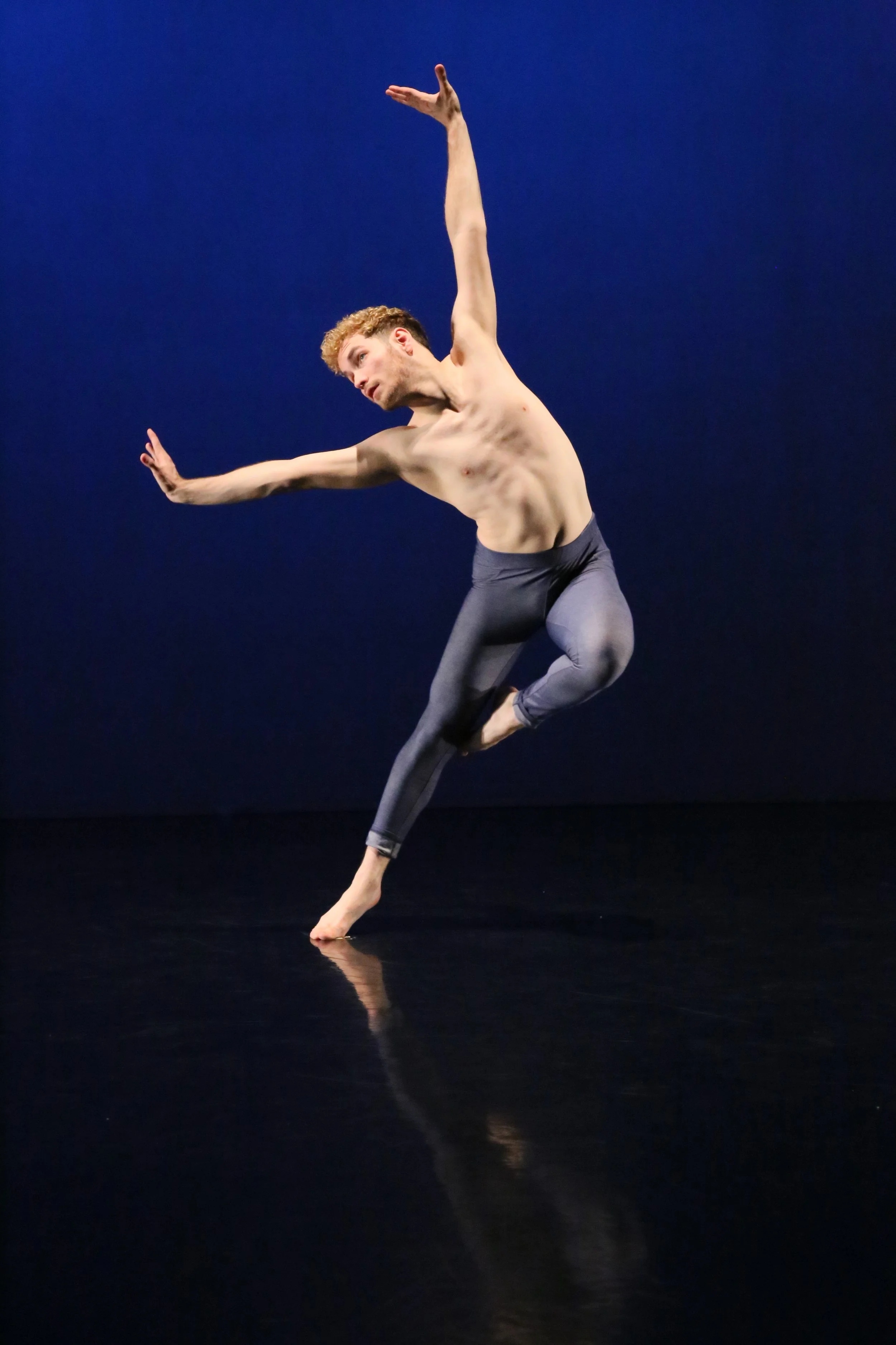 A male dancer performing a contemporary dance move on a dark stage with a dark blue background, wearing gray leggings and no shirt.