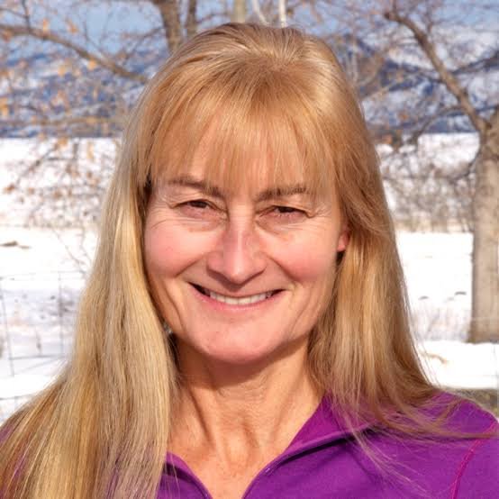 A smiling woman with long blonde hair standing outdoors with snow and trees in the background.