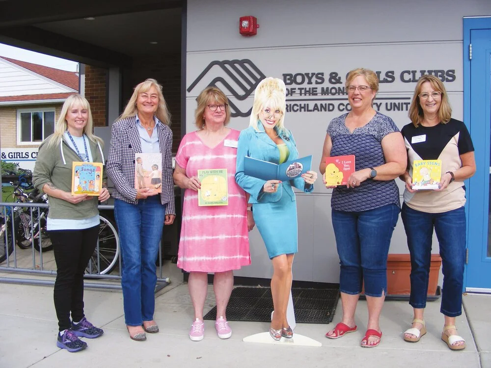 Six women standing in a row outside the Boys & Girls Clubs, holding children's books, with one woman holding a cardboard cutout of a Dolly Parton.