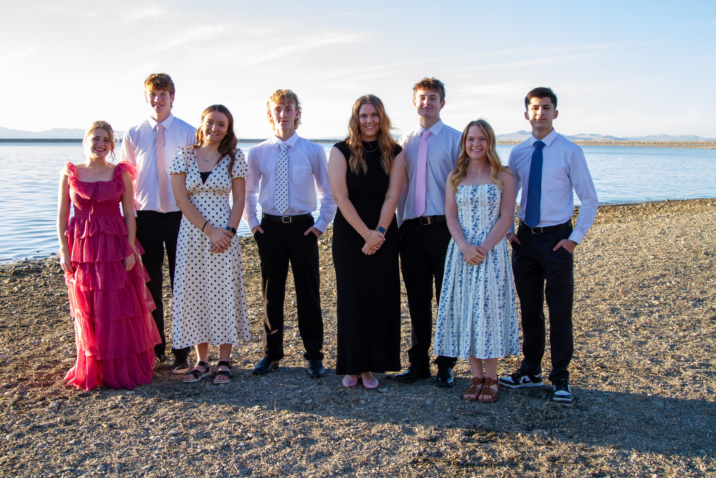 Group of nine young people standing on a rocky beach near a body of water, dressed in semi-formal attire, with mountains in the background.