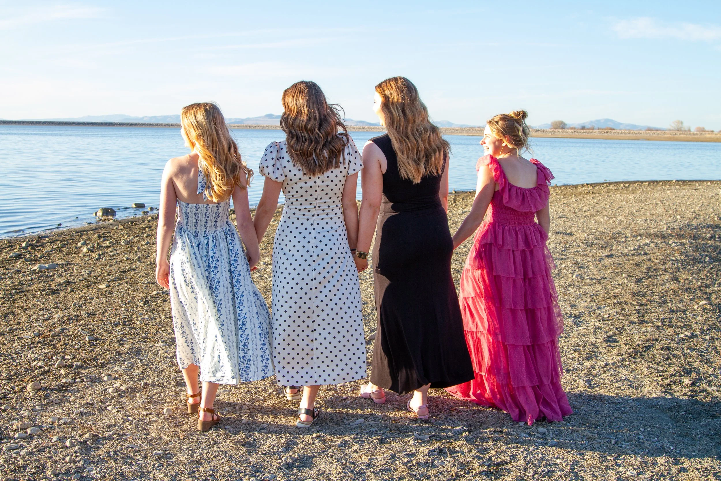Four women in dresses holding hands walk on a beach near a body of water, with mountains visible in the distance.