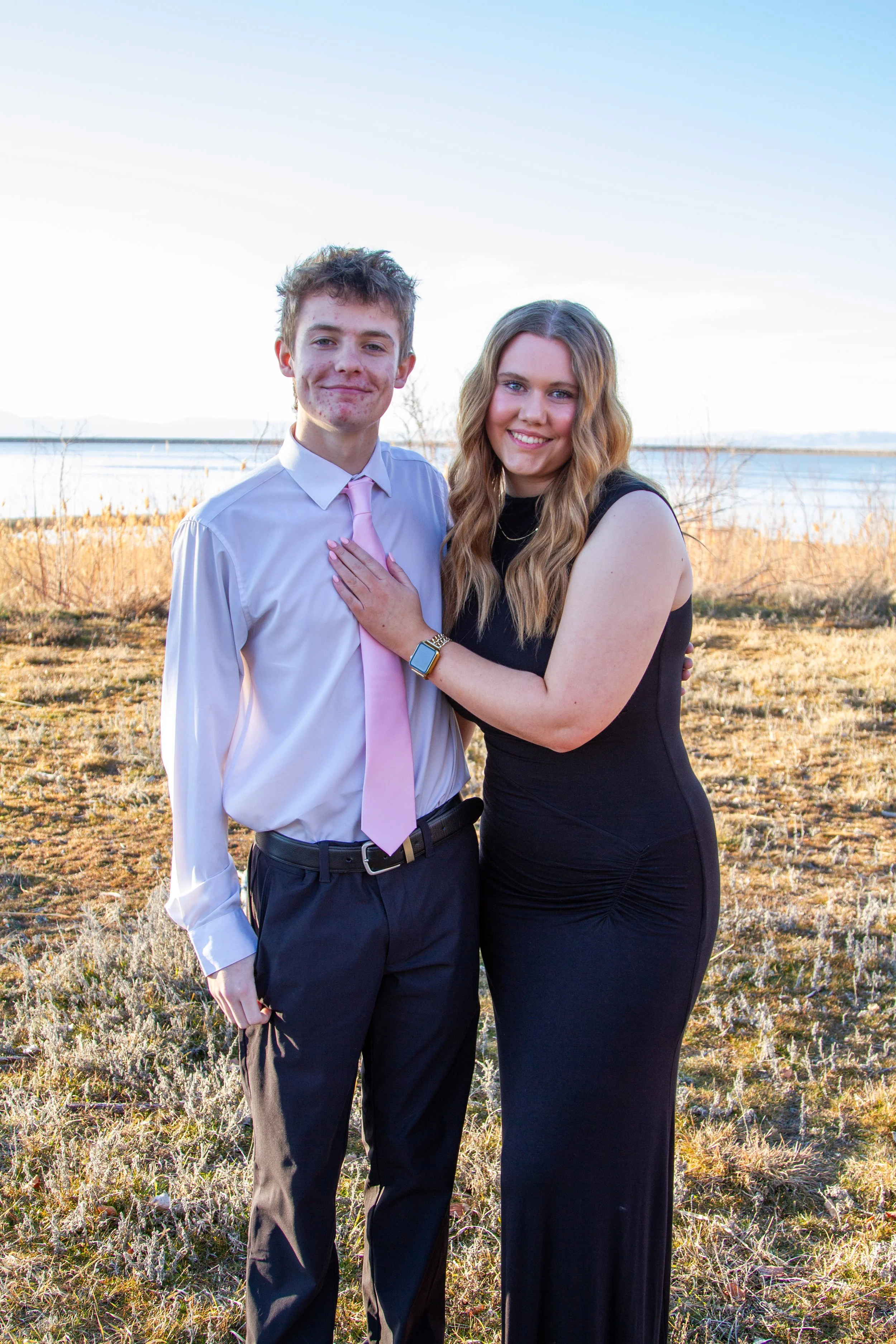 A young man and woman standing outdoors by a body of water, smiling at the camera. The young man is dressed in a white shirt with a pink tie and black pants, while the young woman wears a black sleeveless dress and has long wavy hair.