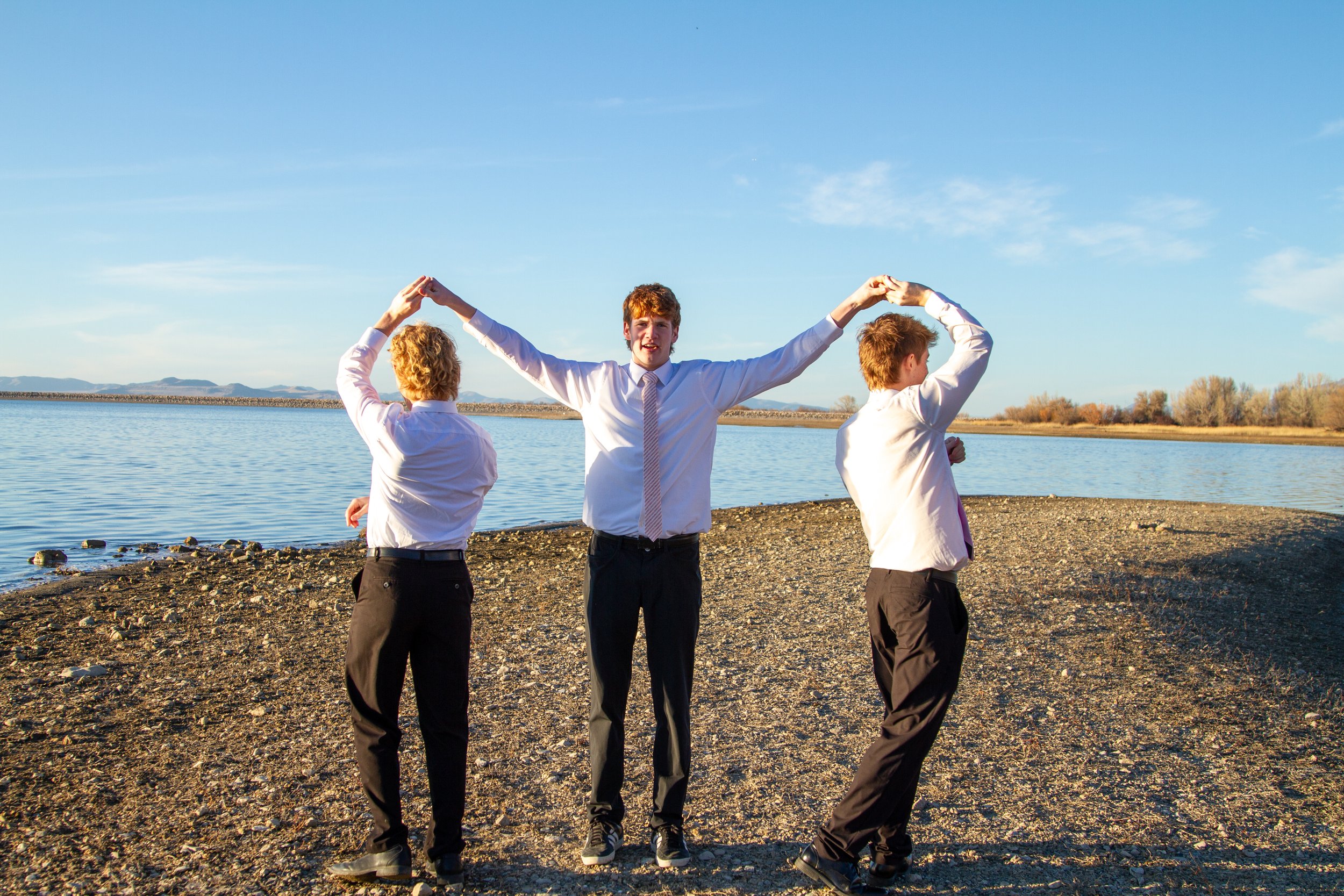 Three young men in formal attire standing by a body of water, with the central figure holding hands with the others above his head, forming an arch.