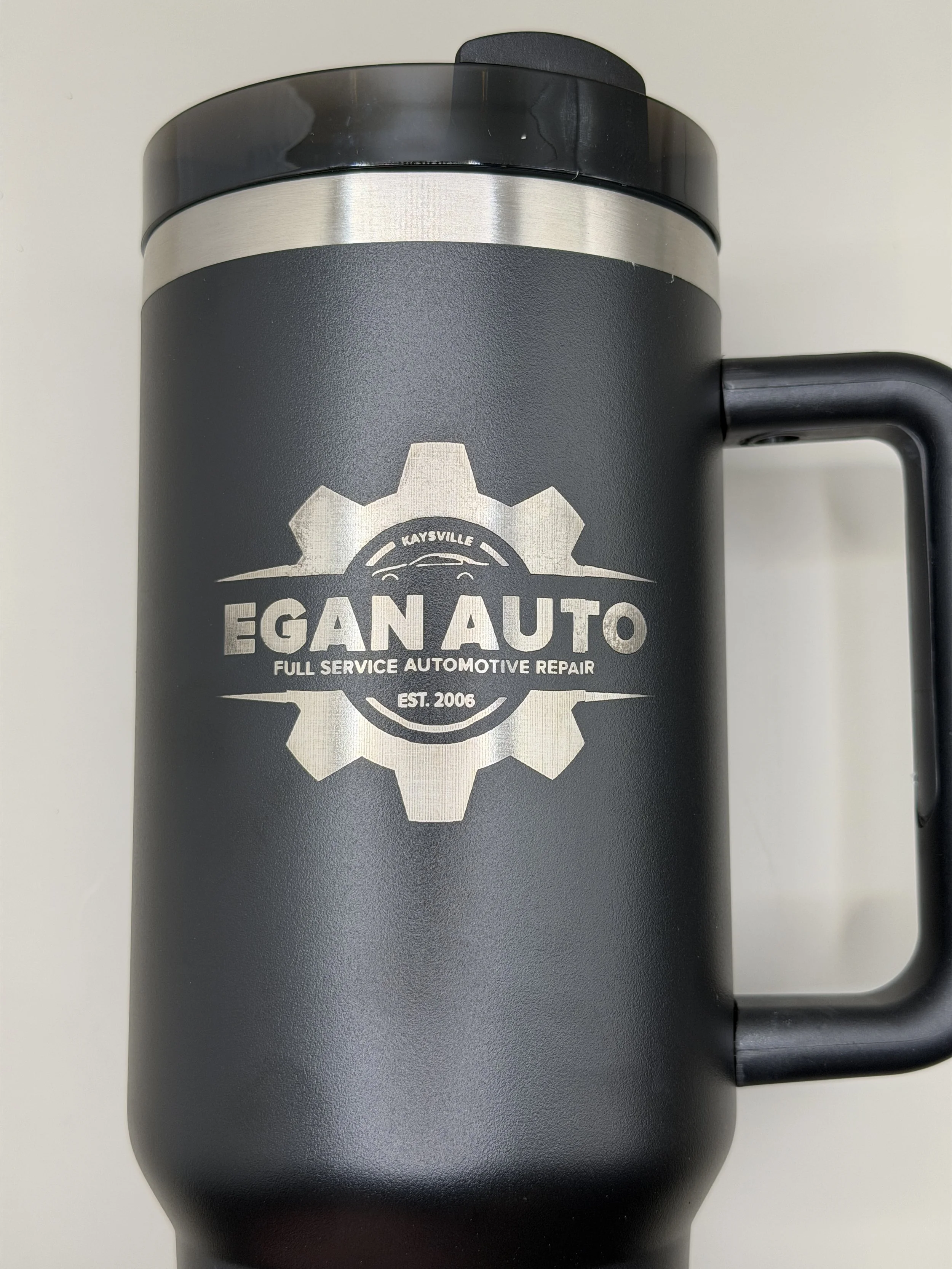 A black travel mug with a silver band and a logo that reads "Egan Auto" and "Full Service Automotive Repair". The mug has a handle on the right side.