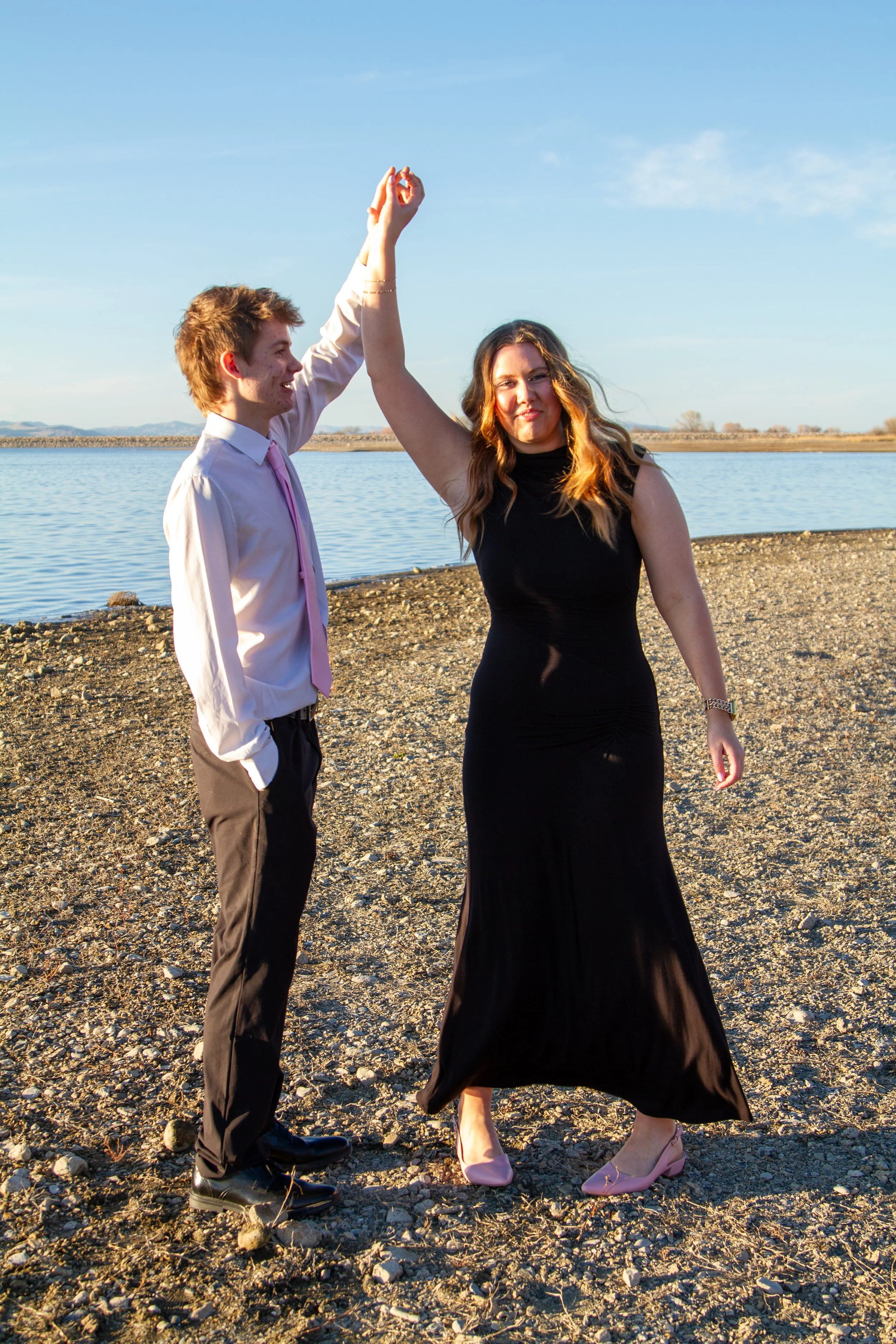 A couple dancing outdoors by a body of water during sunset, with the man in a white shirt and pink tie, and the woman in a black dress and pink shoes, holding hands up in a dance pose.