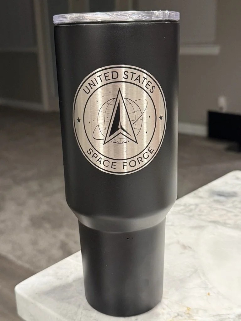 A black insulated tumbler with a silver emblem of the United States Space Force logo on it, sitting on a white surface in a room with grey carpet and walls.