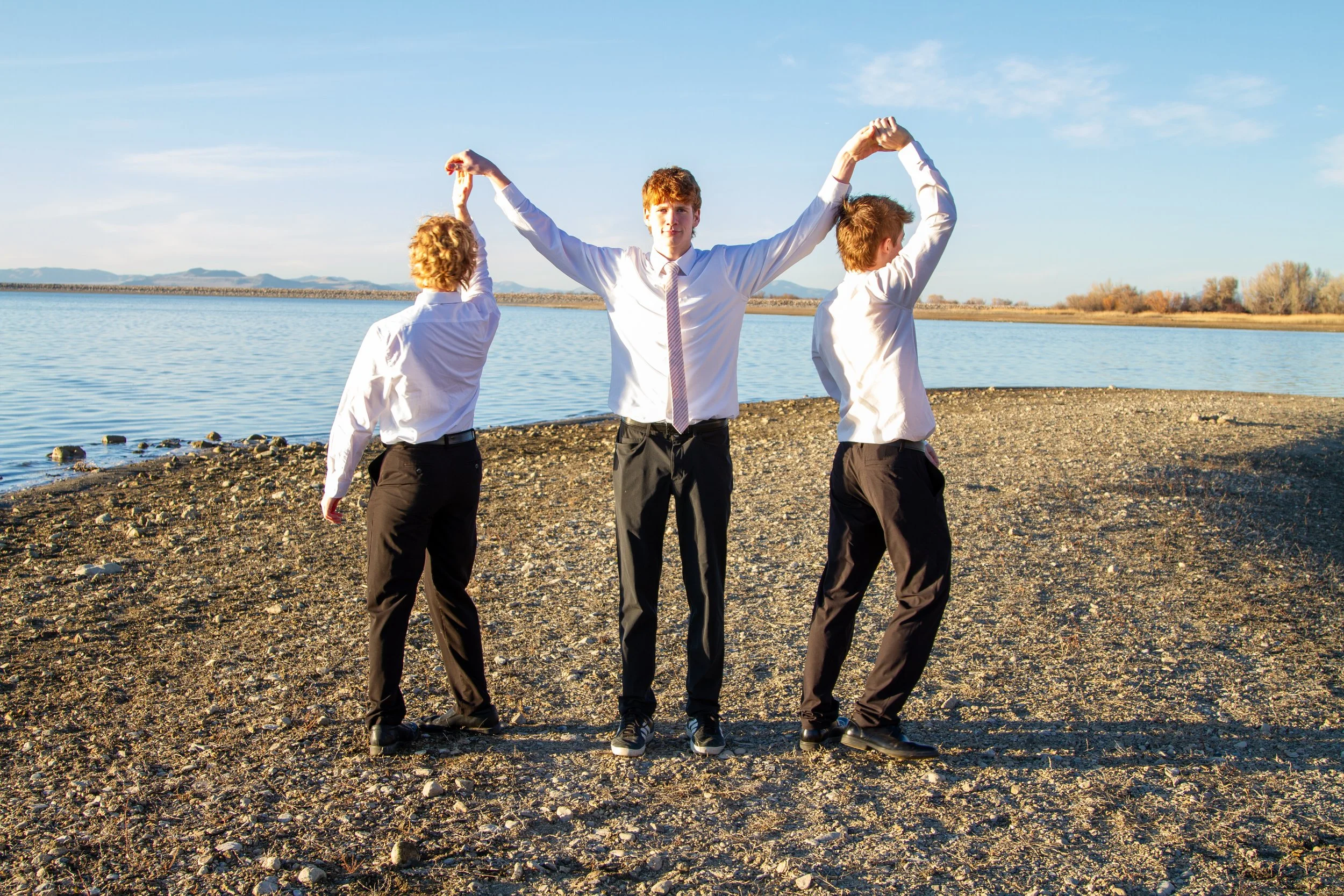 Three young men in business suits holding hands with their arms raised, standing on a rocky shoreline near a body of water during daytime.
