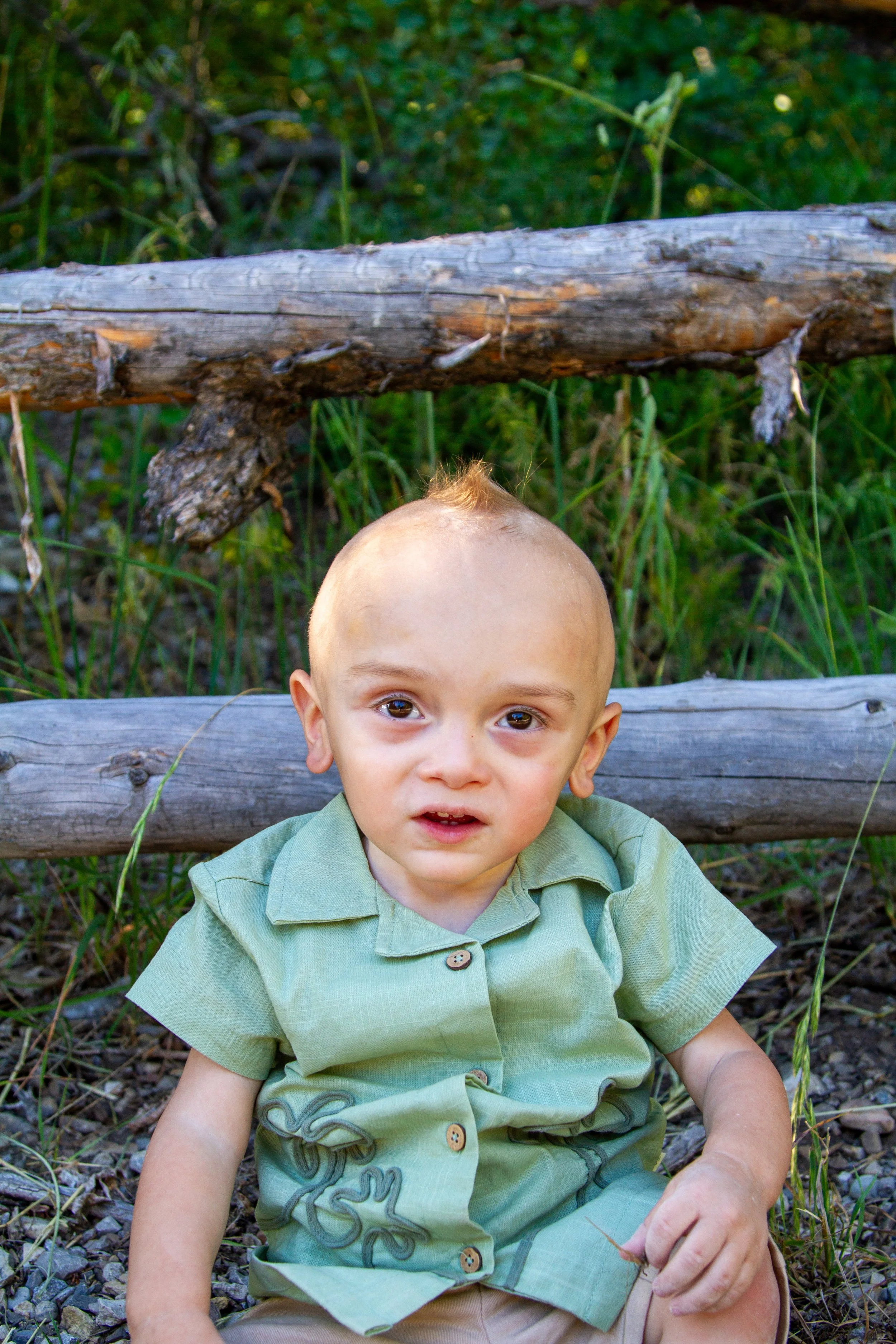 Young boy with light skin and short light-colored hair sitting outdoors on a gravelly ground, wearing a light green shirt and khaki shorts, with a wooden fence and green foliage in the background.