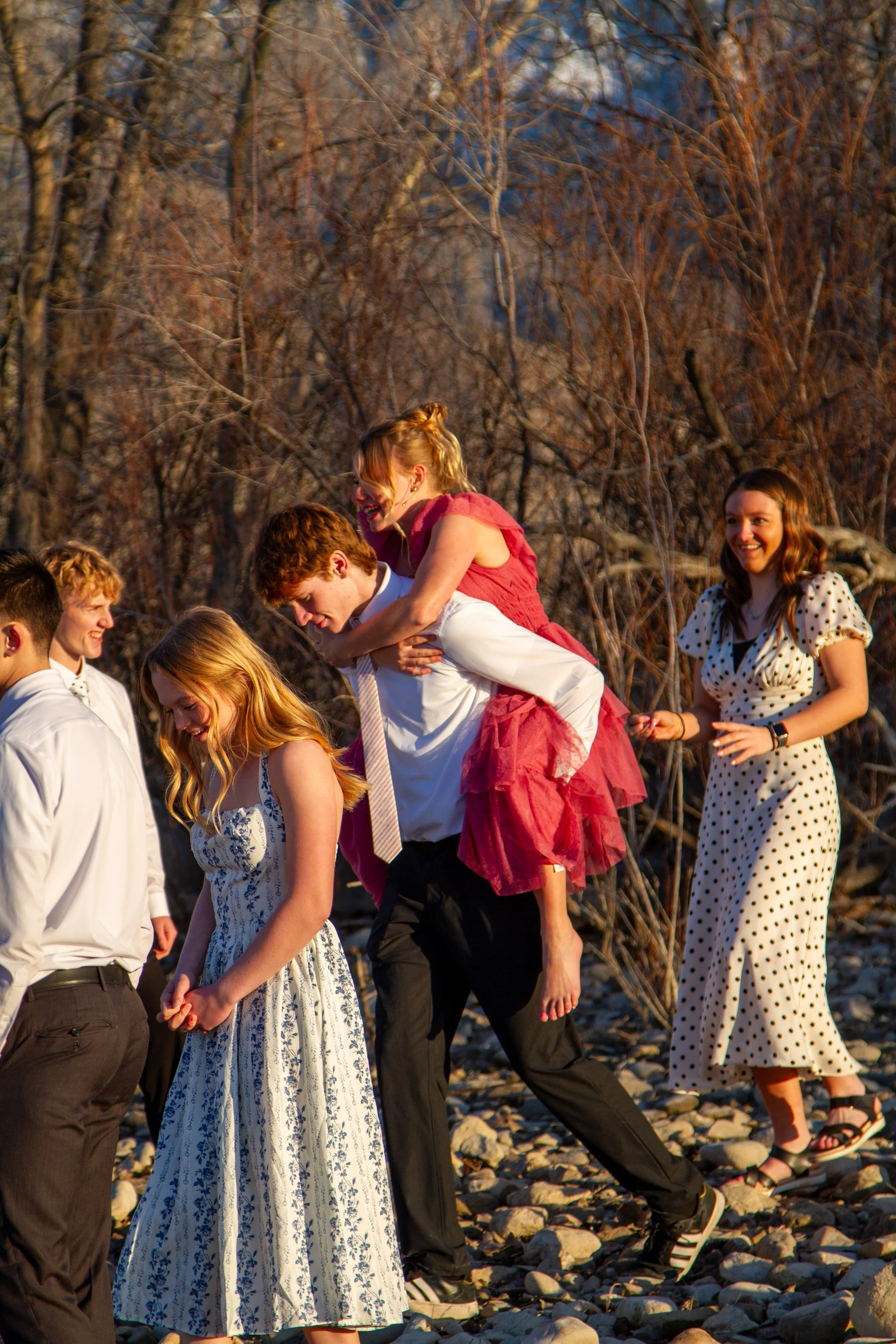 Group of young people outdoors, with some smiling, enjoying a playful moment on a rocky ground during sunset, with barren trees in the background.