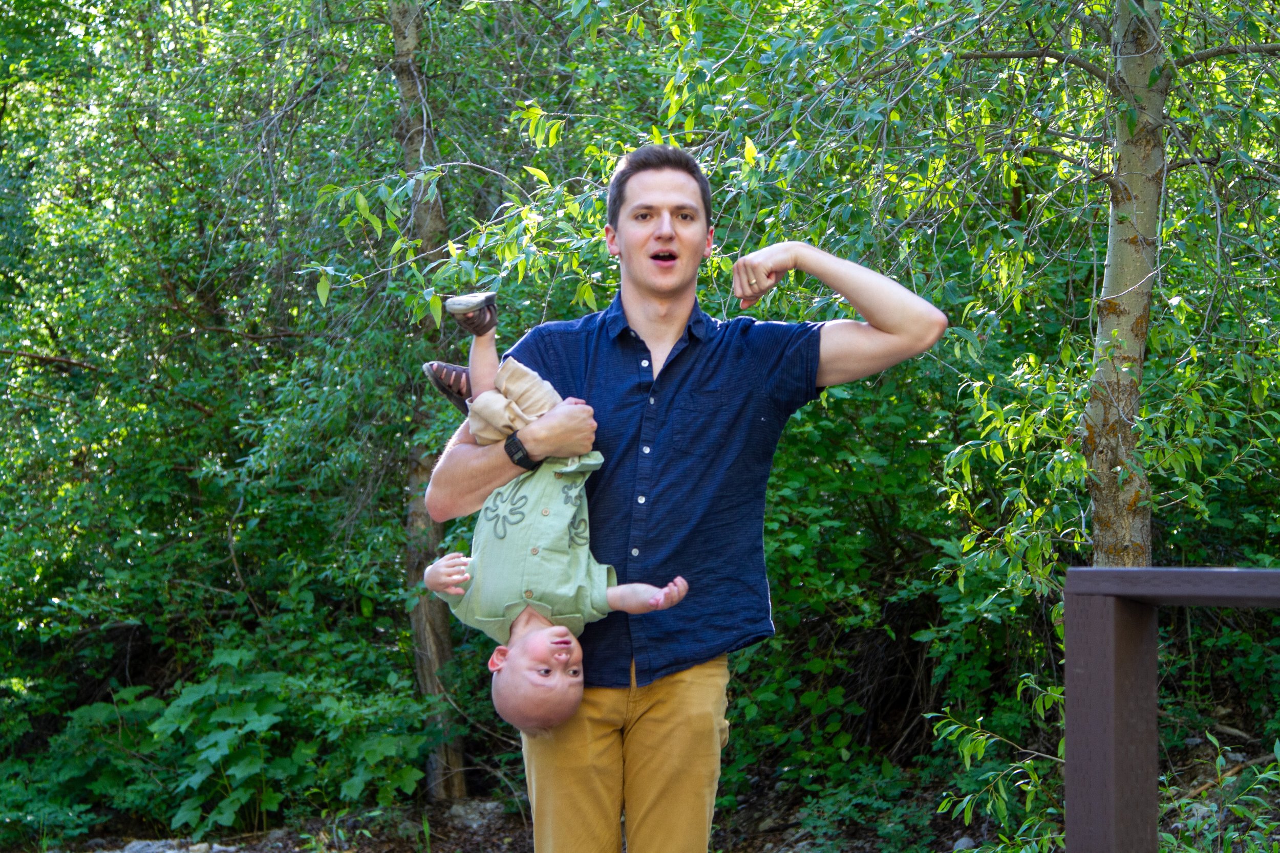 A man standing outdoors in front of green trees, with a child upside down hanging from his shoulders, and another child clutching his neck. The man is wearing a dark blue shirt and yellow pants, while the children are in light-colored clothing. The s