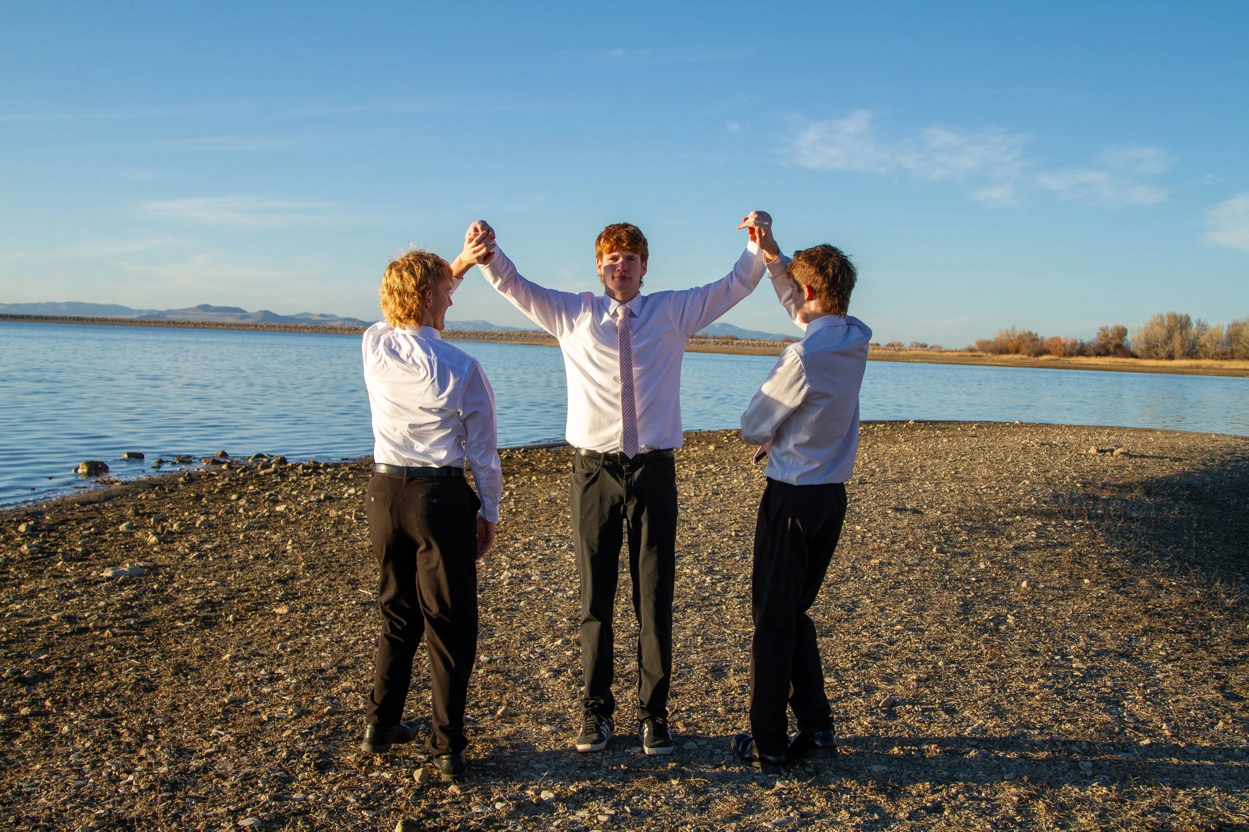 Three young men in business attire standing on a sandy shoreline near a body of water during sunset, with mountains in the background.