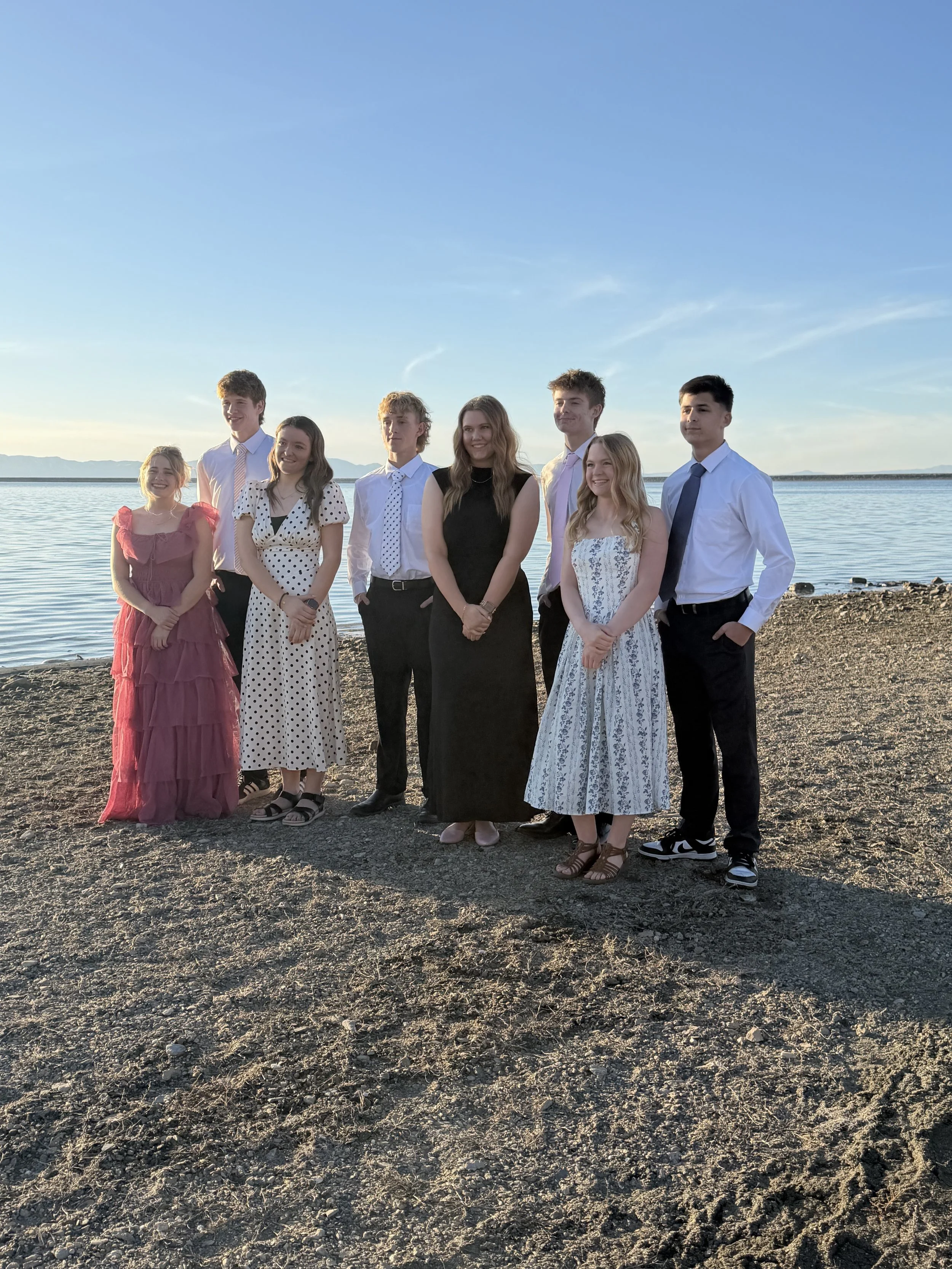 A group of eight young people standing on a rocky beach near a body of water during sunset, dressed in formal or semi-formal attire.