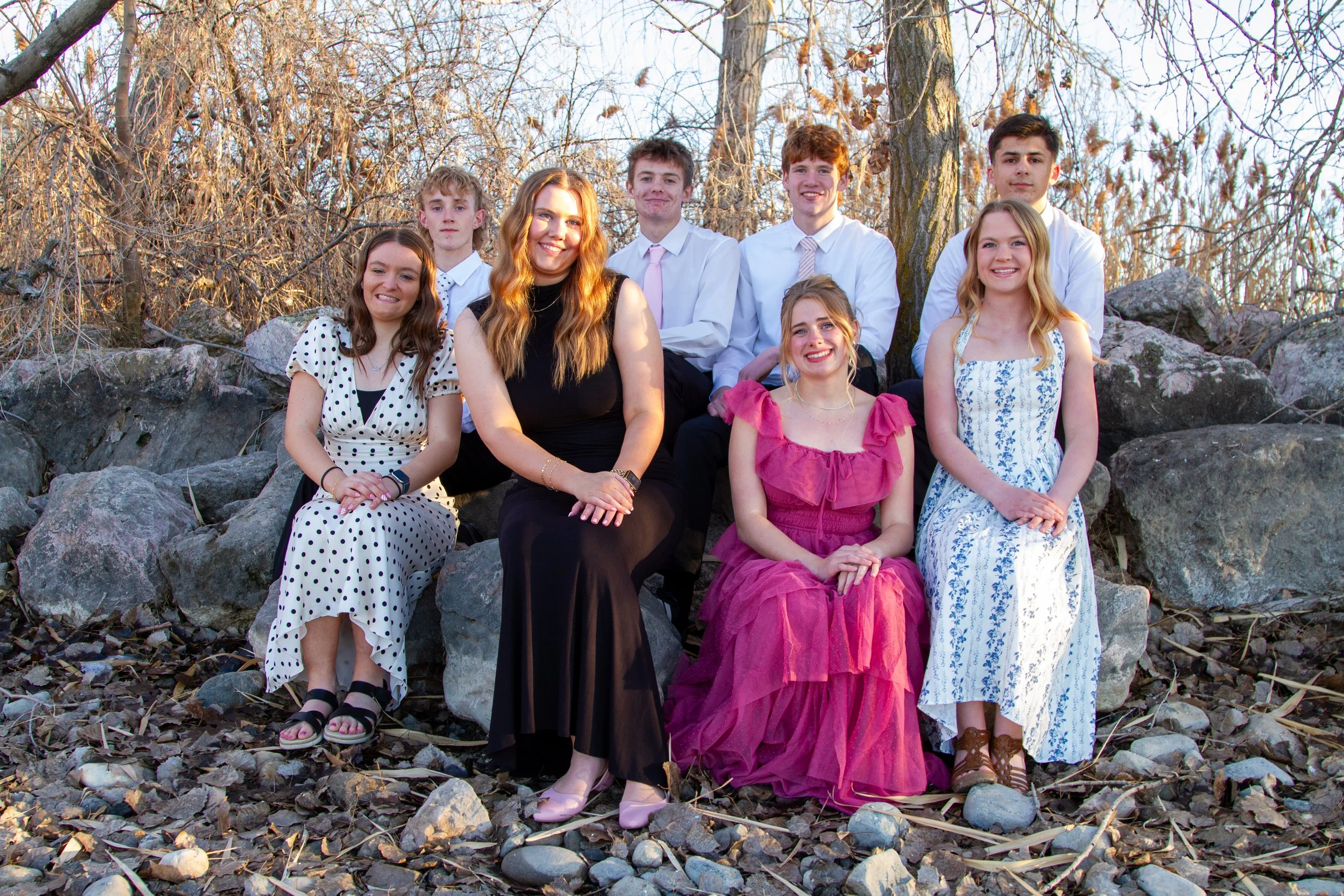 Group of nine young people, four women and five men, sitting on rocks outdoors during sunset, smiling at the camera, with leafless trees in the background.