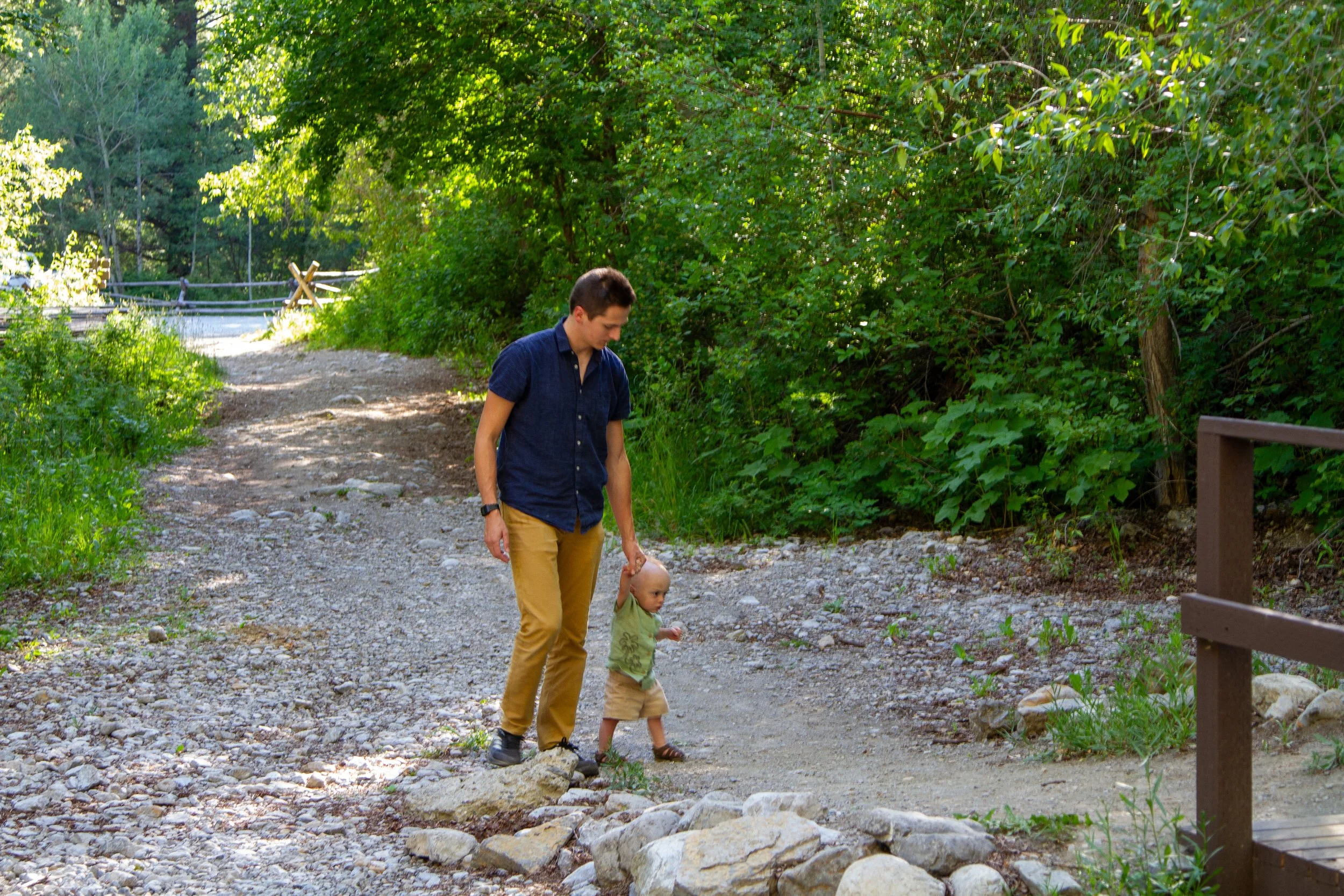 A man and a young child walking together on a gravel path surrounded by green trees during daytime.