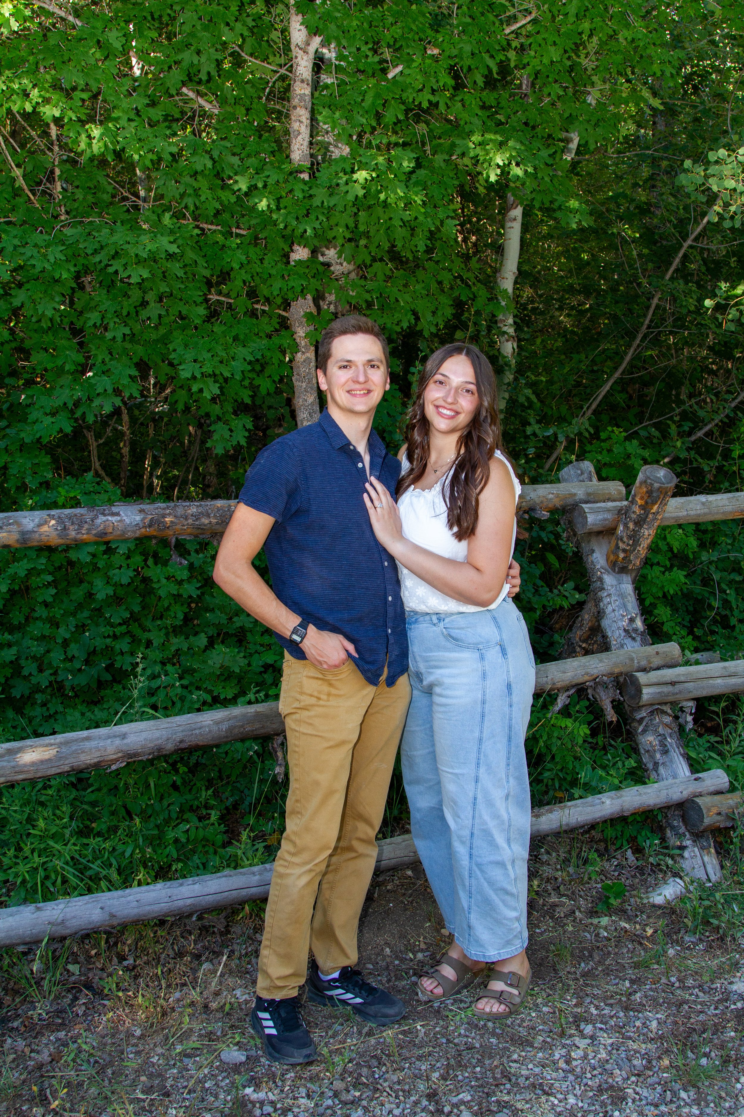 A young man and woman stand close together outdoors in front of a wooded area with green leaves, smiling at the camera. The man is wearing a navy blue shirt and tan pants, and the woman is wearing a white blouse and light blue jeans. They are standin