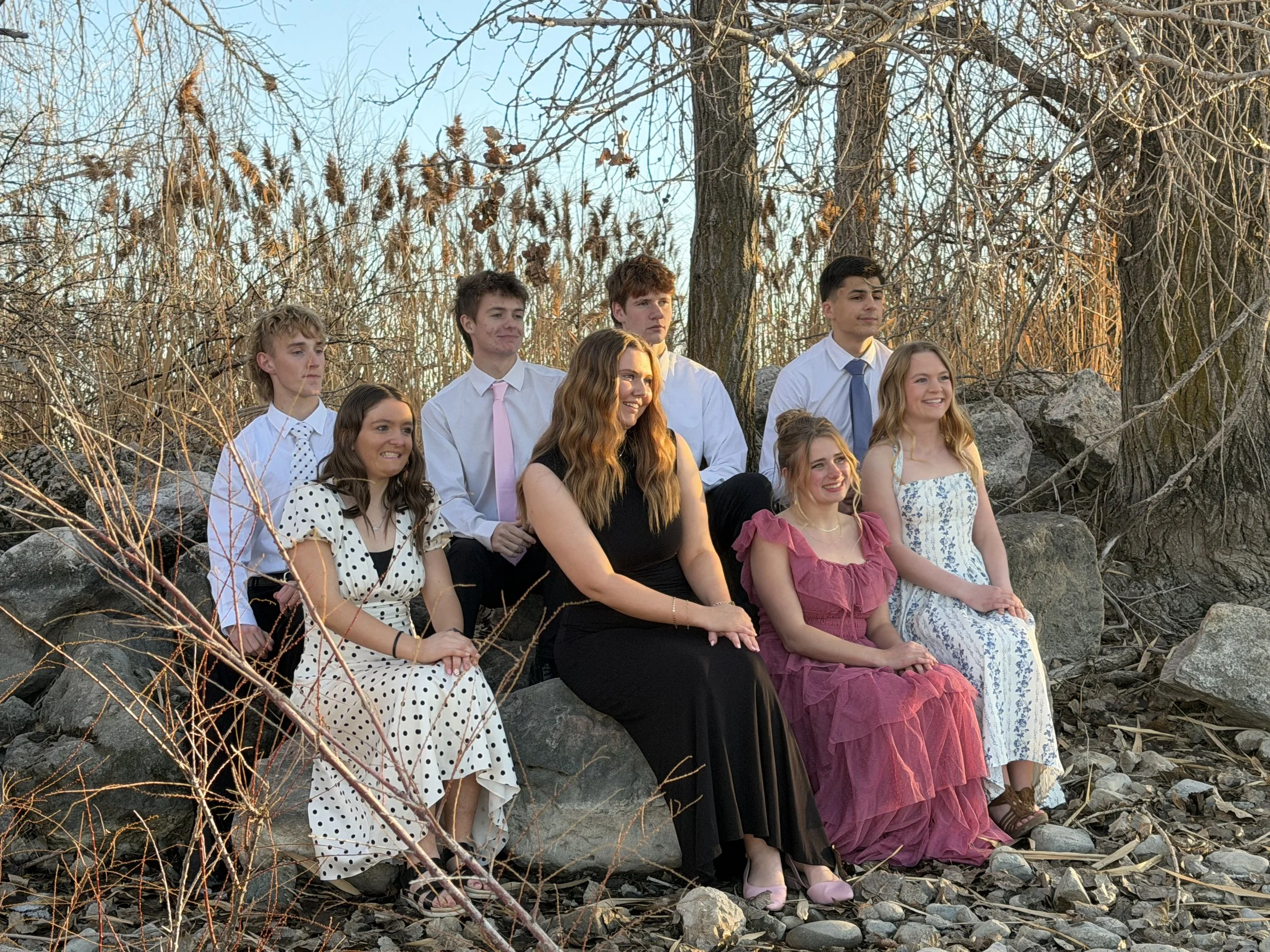 A group of nine young people sitting on rocks outdoors during sunset, dressed in semi-formal attire, with trees and dry branches in the background.