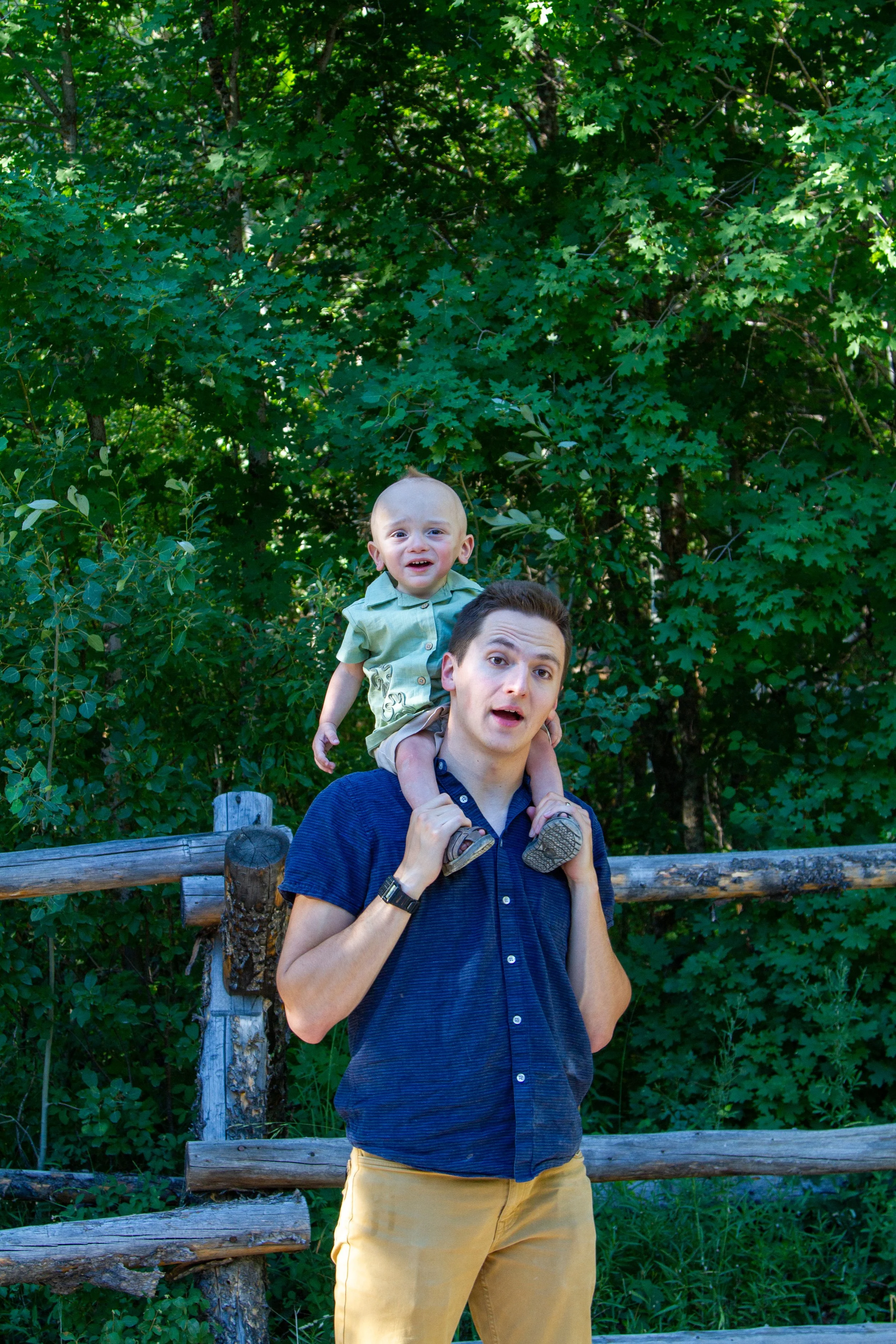 A man with a young boy on his shoulders, in front of a green, leafy background.