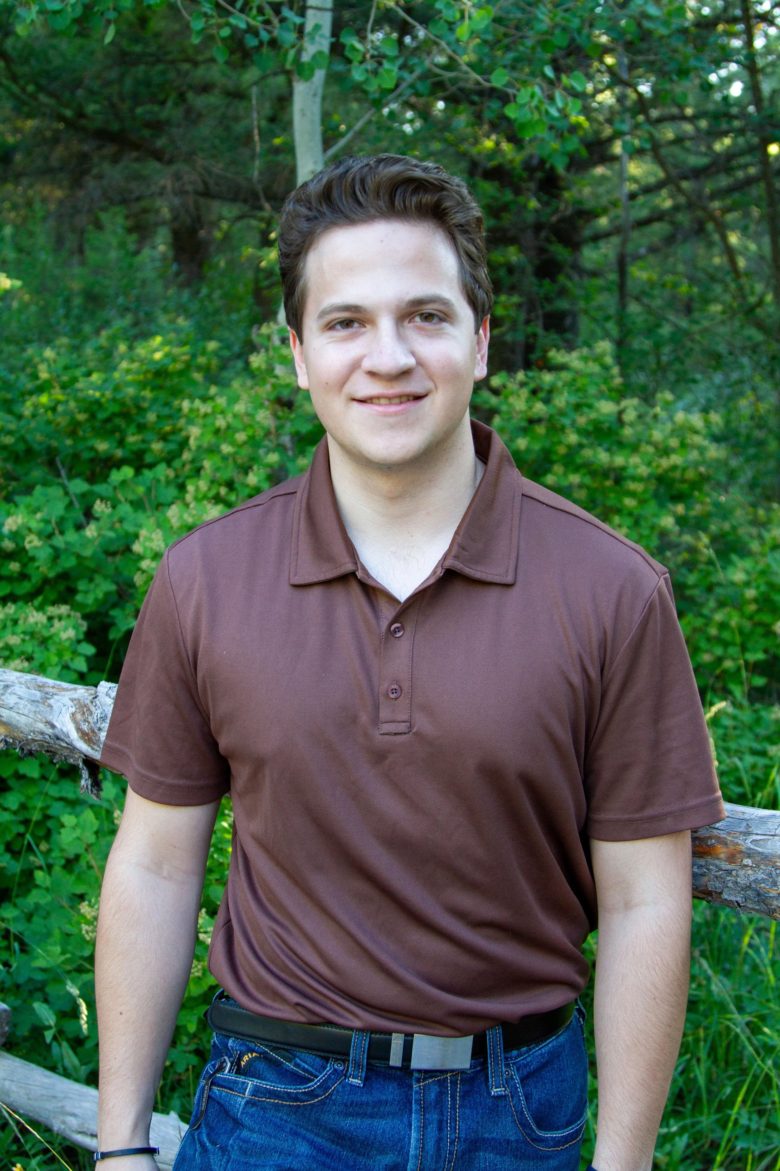 A young man with short dark hair, wearing a brown polo shirt and blue jeans, standing outdoors in front of green trees and bushes, smiling at the camera.