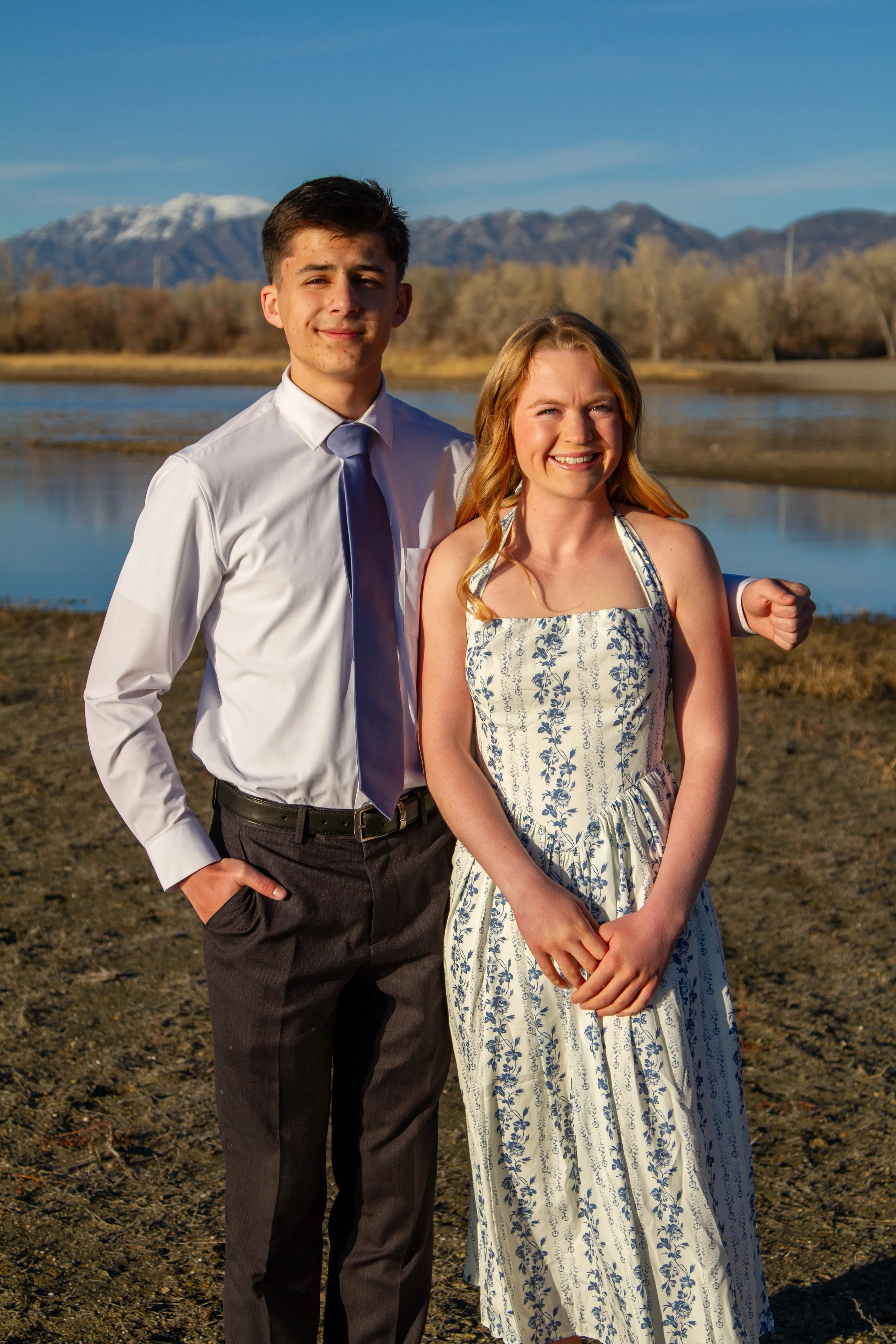 A young man and woman standing outdoors near a body of water with mountains in the background, smiling at the camera.