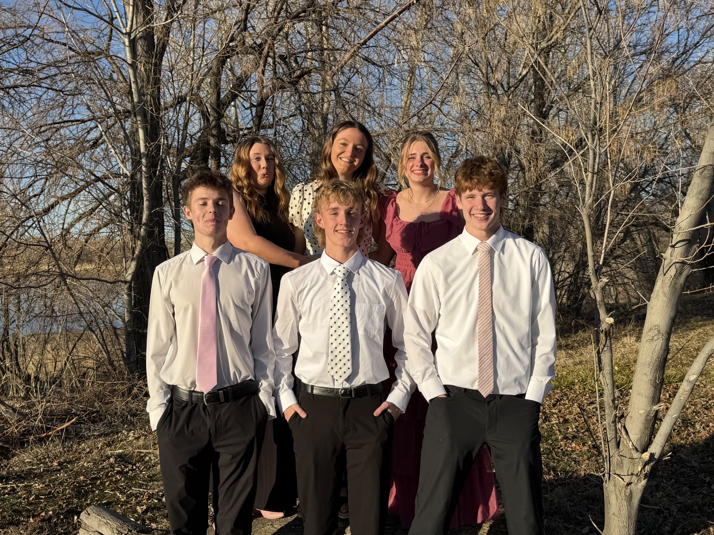 Group of six teenagers, three boys and three girls, standing outdoors in front of leafless trees during daytime with clear sky, dressed in semi-formal attire, smiling for a photo.