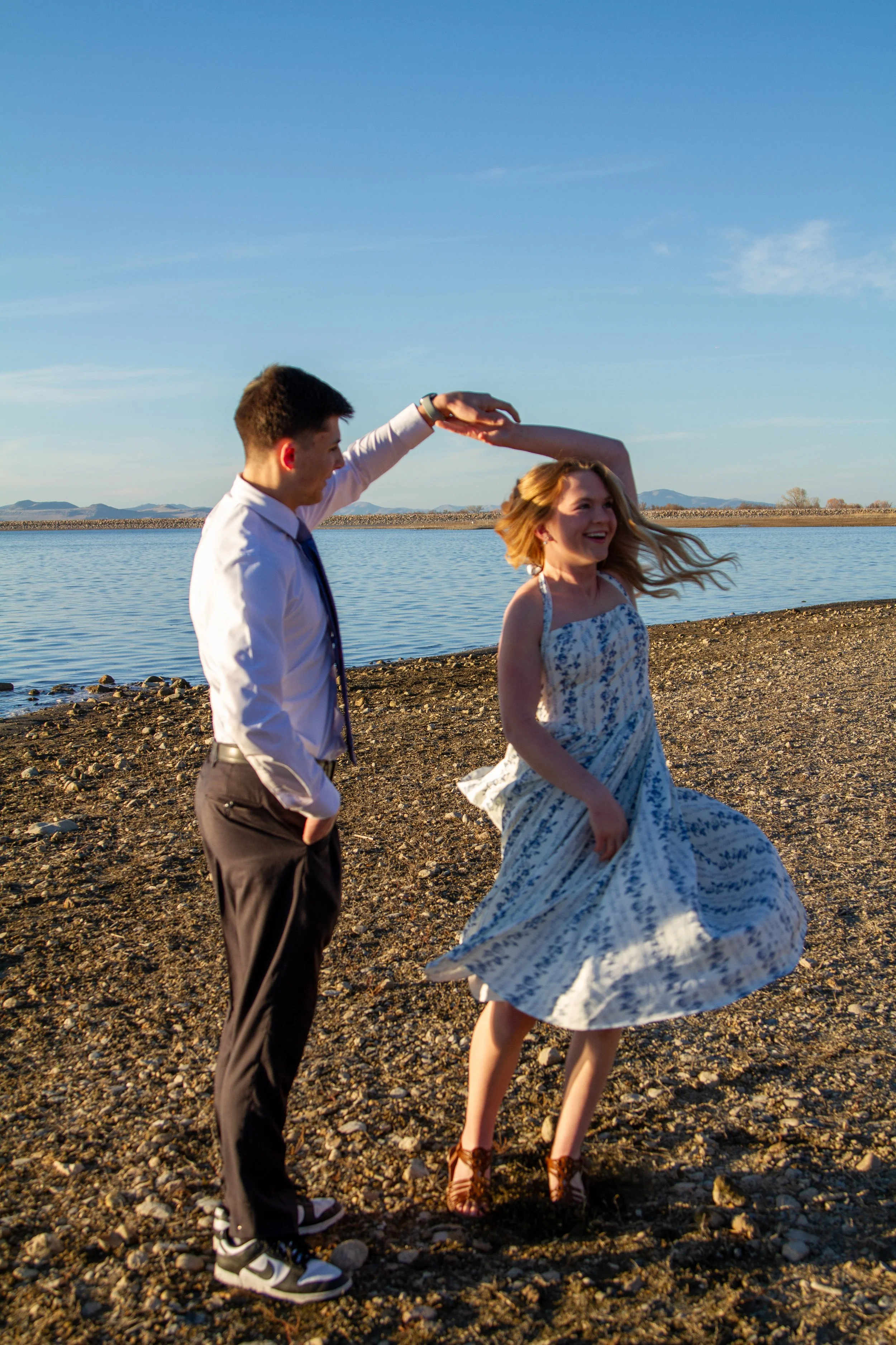A young man and woman are dancing on a rocky beach by the water during sunset, with the man twirling the woman.