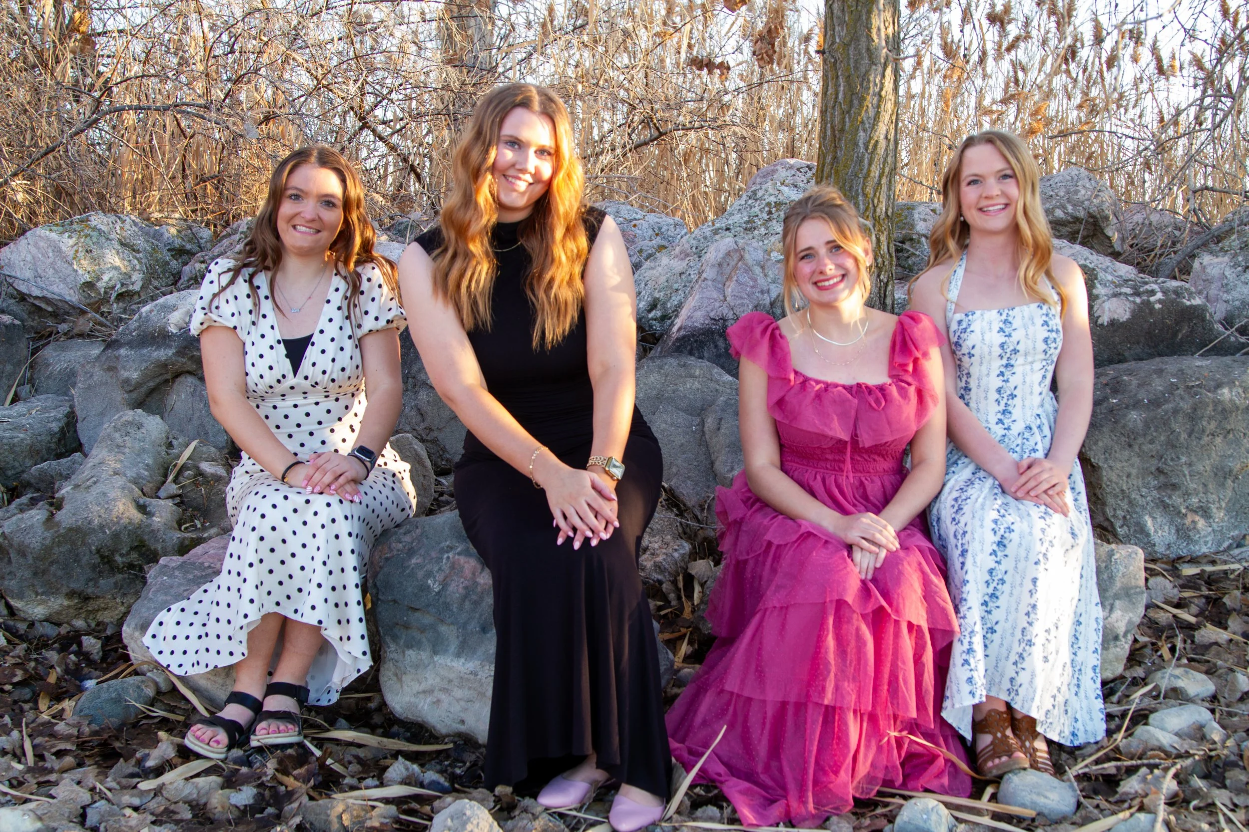 Four young women sitting on rocks outdoors during daylight, with trees and dry foliage in the background. They are smiling and dressed in summer or spring dresses.