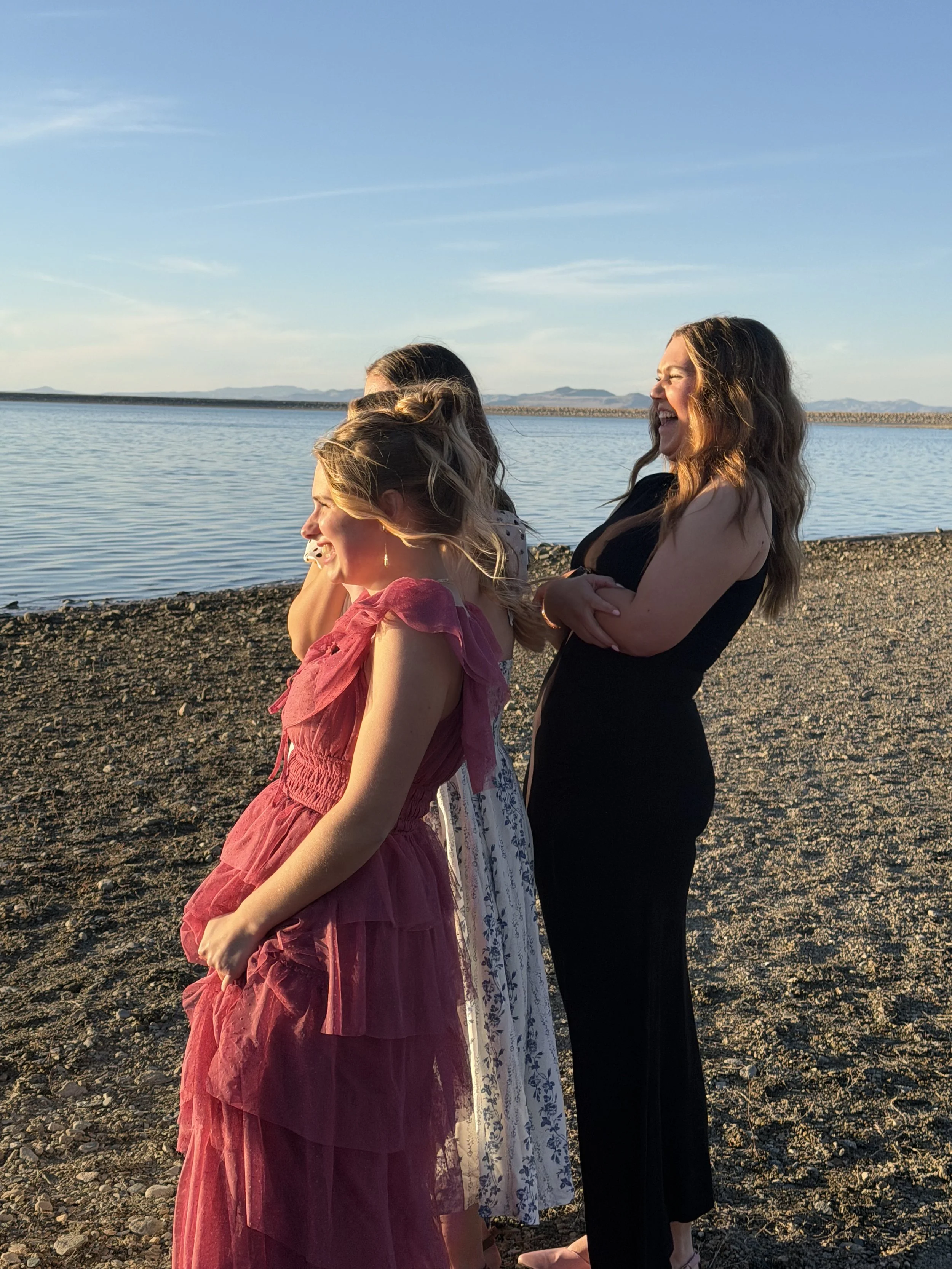 Four women standing on a rocky beach near water, laughing and enjoying the sunset.