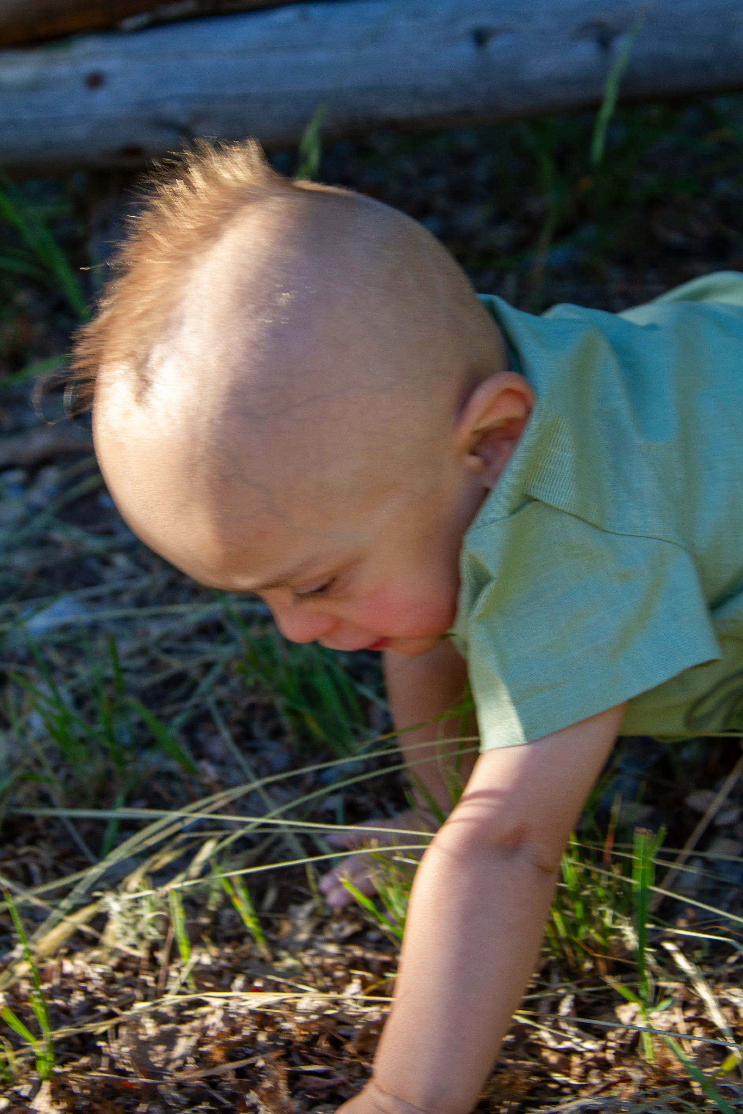 A young child with a Mohawk haircut crawling outdoors among grass and dirt, wearing a light green shirt.