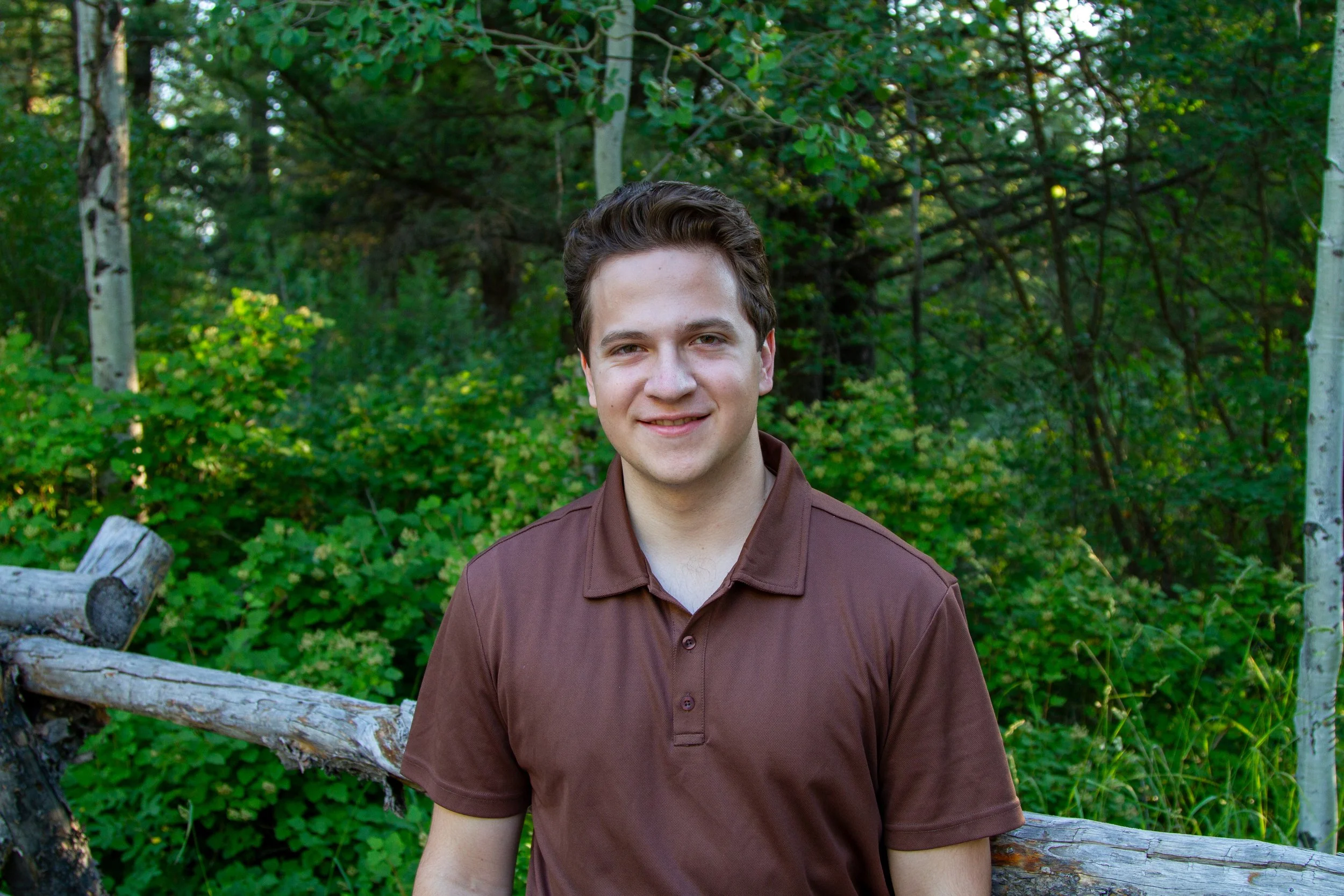 A young man with brown hair wearing a brown shirt standing outdoors in a wooded area with green foliage and trees, smiling at the camera.
