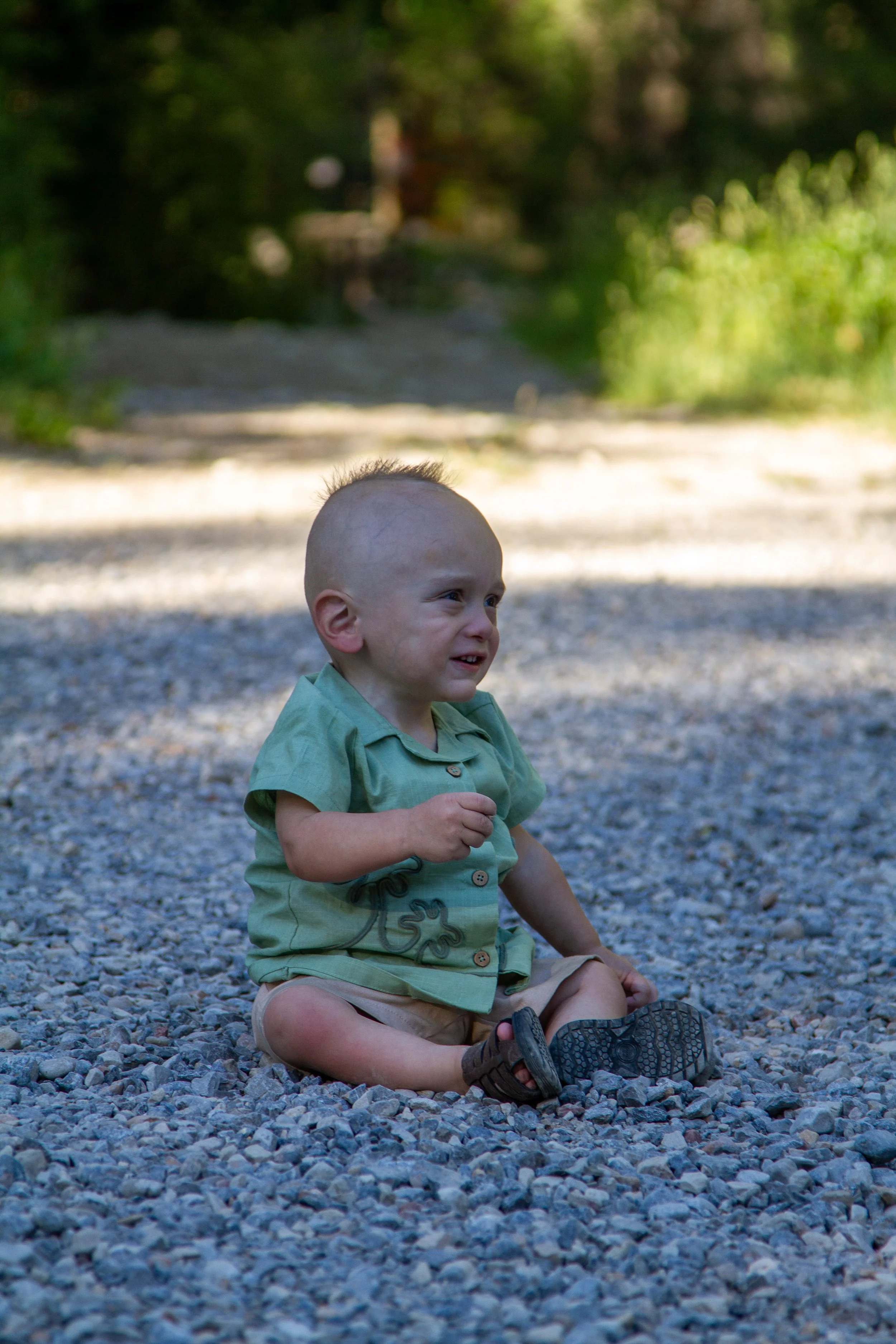 A young child with a shaved head sitting on a gravel surface outdoors, wearing a green shirt and beige shorts, with a blurred natural background of trees and sunlight.