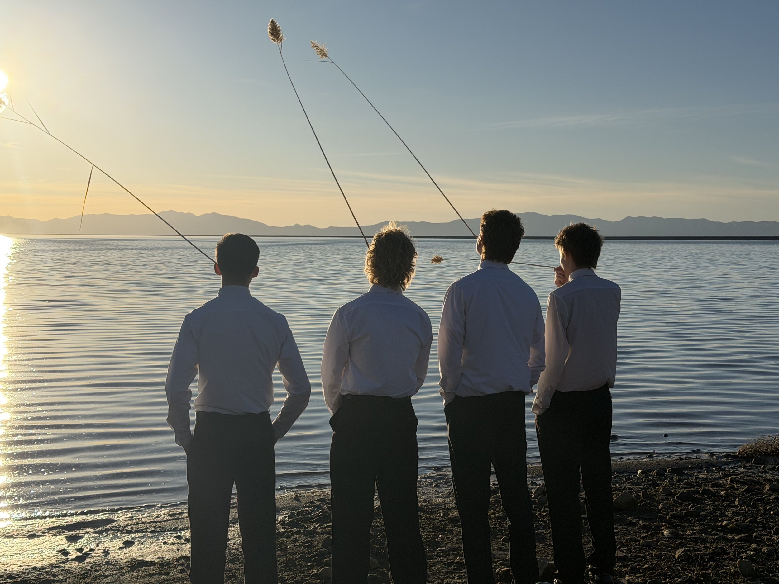 Four men stand on a rocky shoreline, facing water with mountains in the distance, as the sun sets. They are dressed in white shirts and dark trousers, some with hands in pockets, one with hand on chin, and two fishing rods are casting lines into the 
