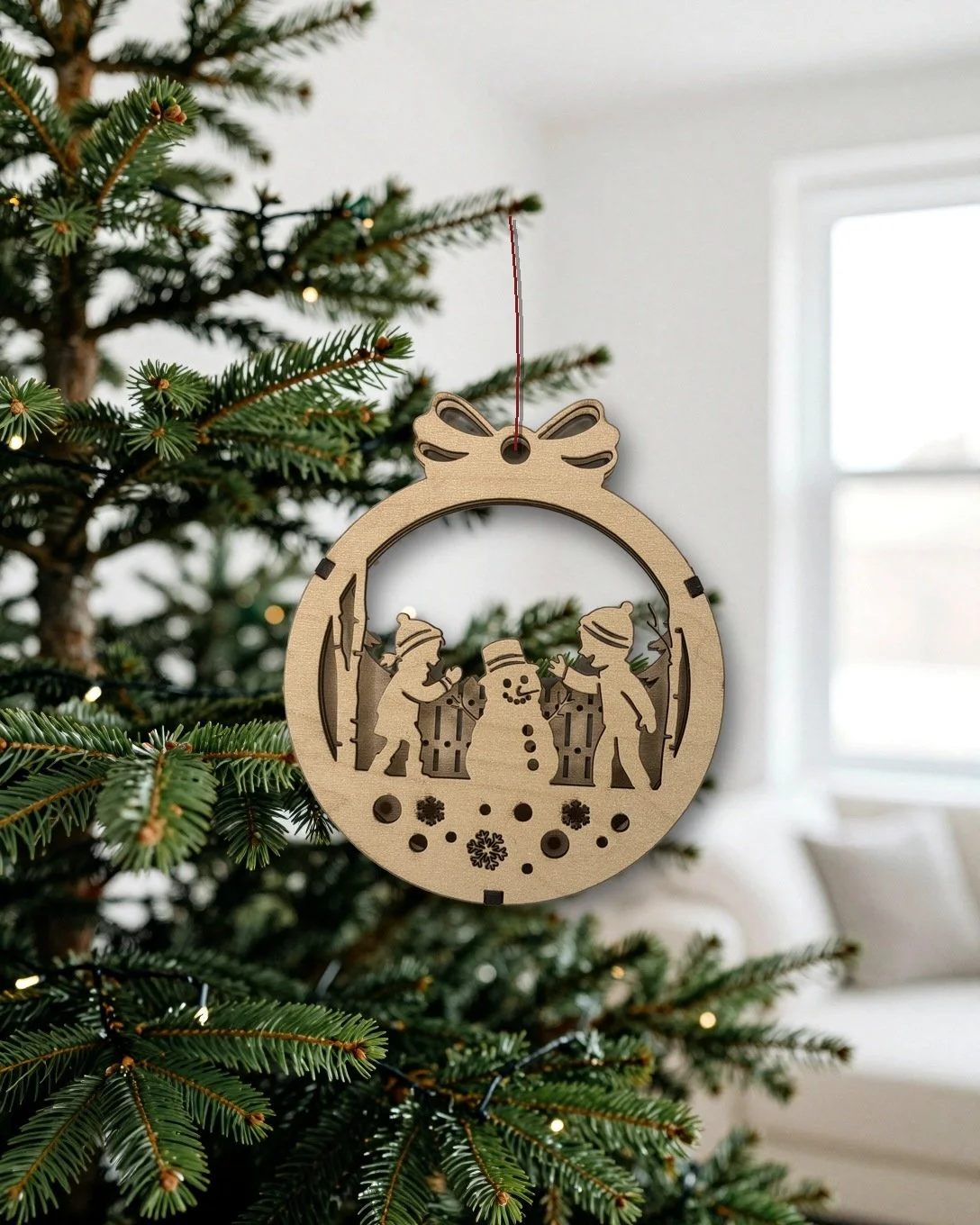 Maple kids building a snowman layered wood Christmas ornament hanging on a tree in a bright living room