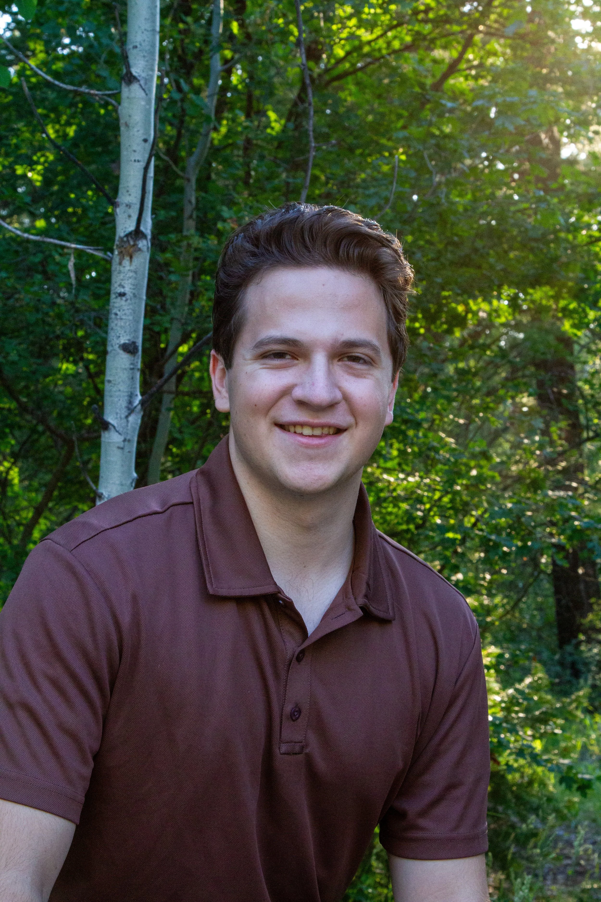 A young man with dark, wavy hair and a light complexion smiling outdoors. He is wearing a maroon polo shirt. The background features green trees with sunlight filtering through the leaves.