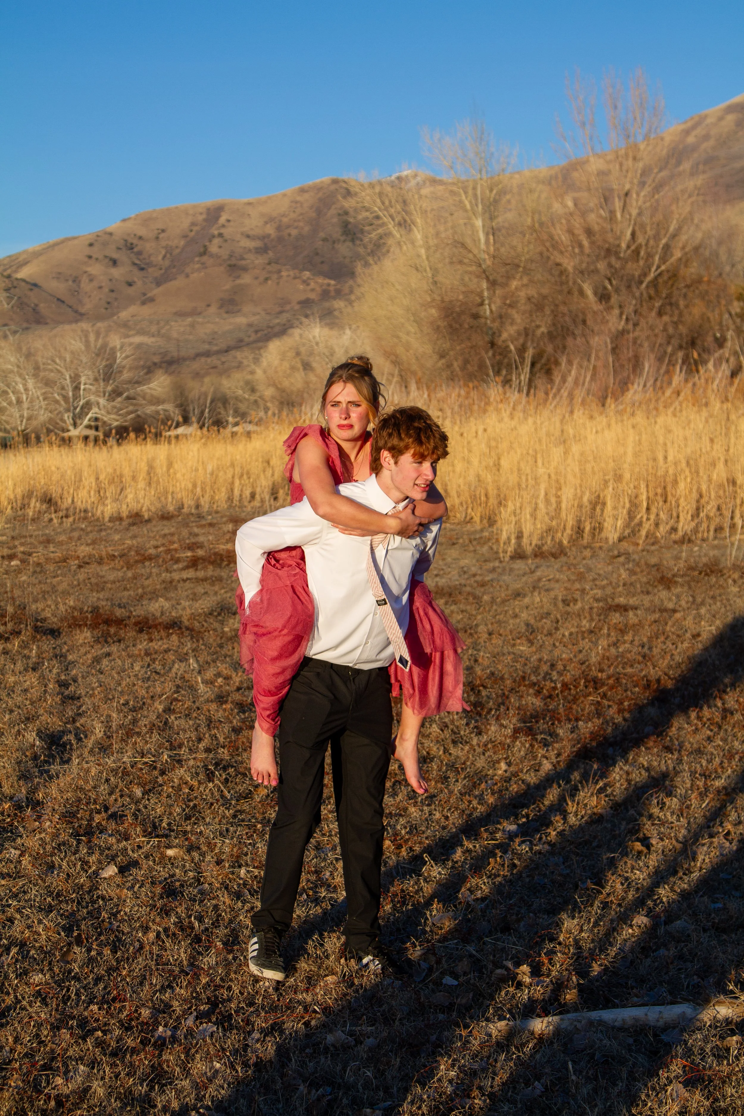 A young man in a white shirt and black pants carries a young girl in a pink dress on his back in a rural outdoor setting with dry grass, trees, and mountains in the background.