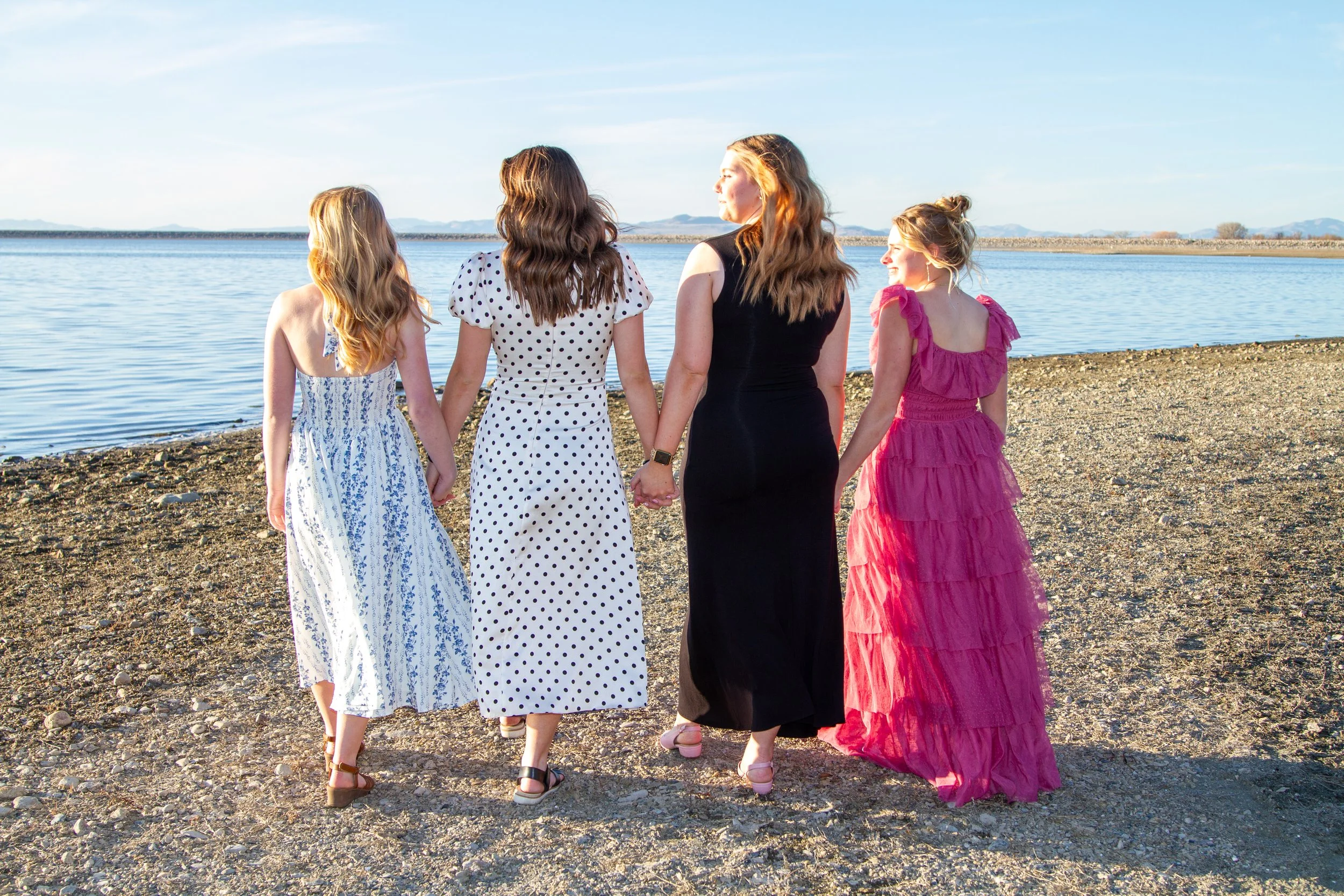 Four women in colorful dresses holding hands and walking on a rocky beach near a lake with mountains in the background.