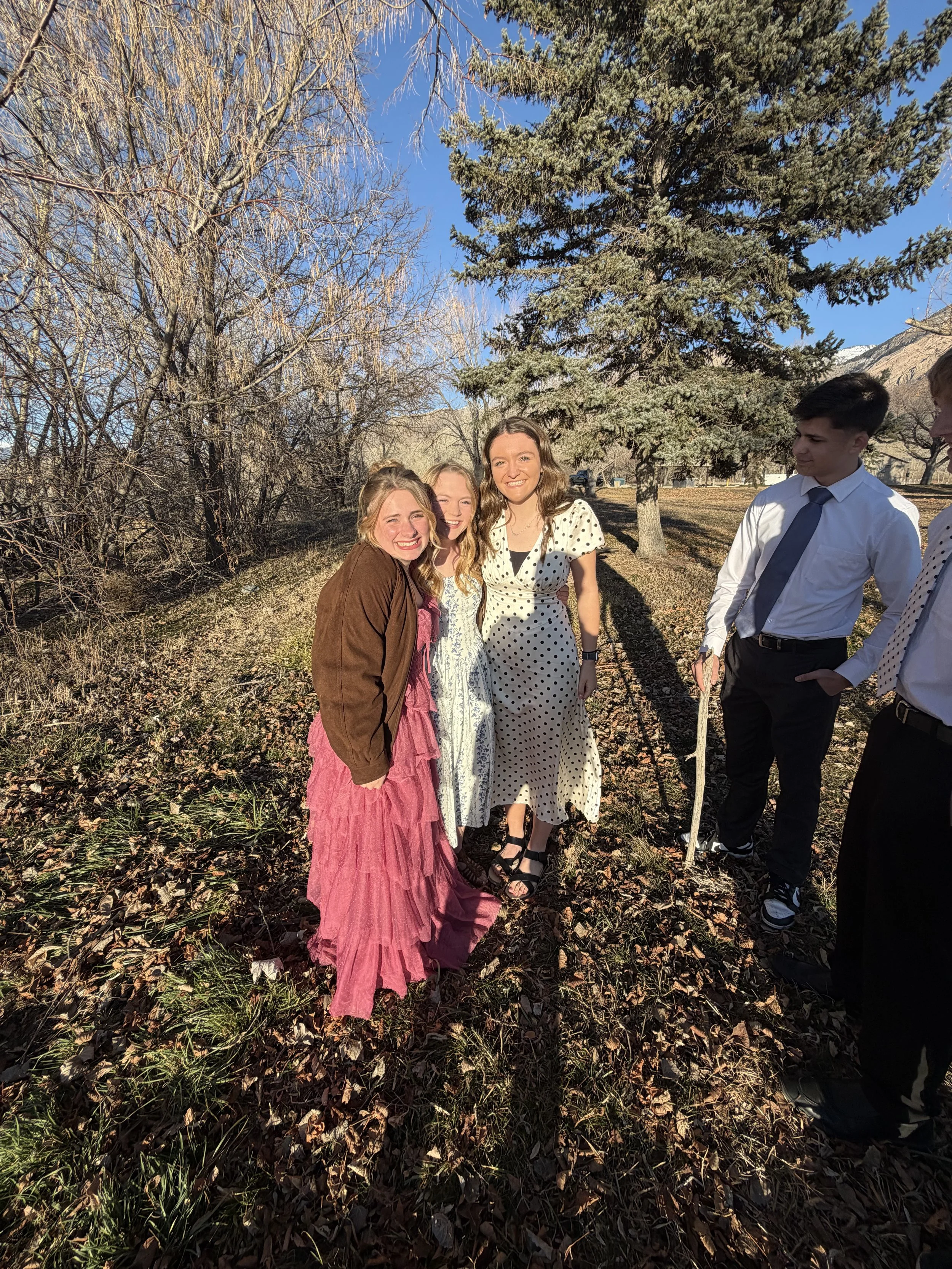 Group of five young people outdoors on a sunny day, standing on a wooded path, smiling and dressed in semi-formal attire.