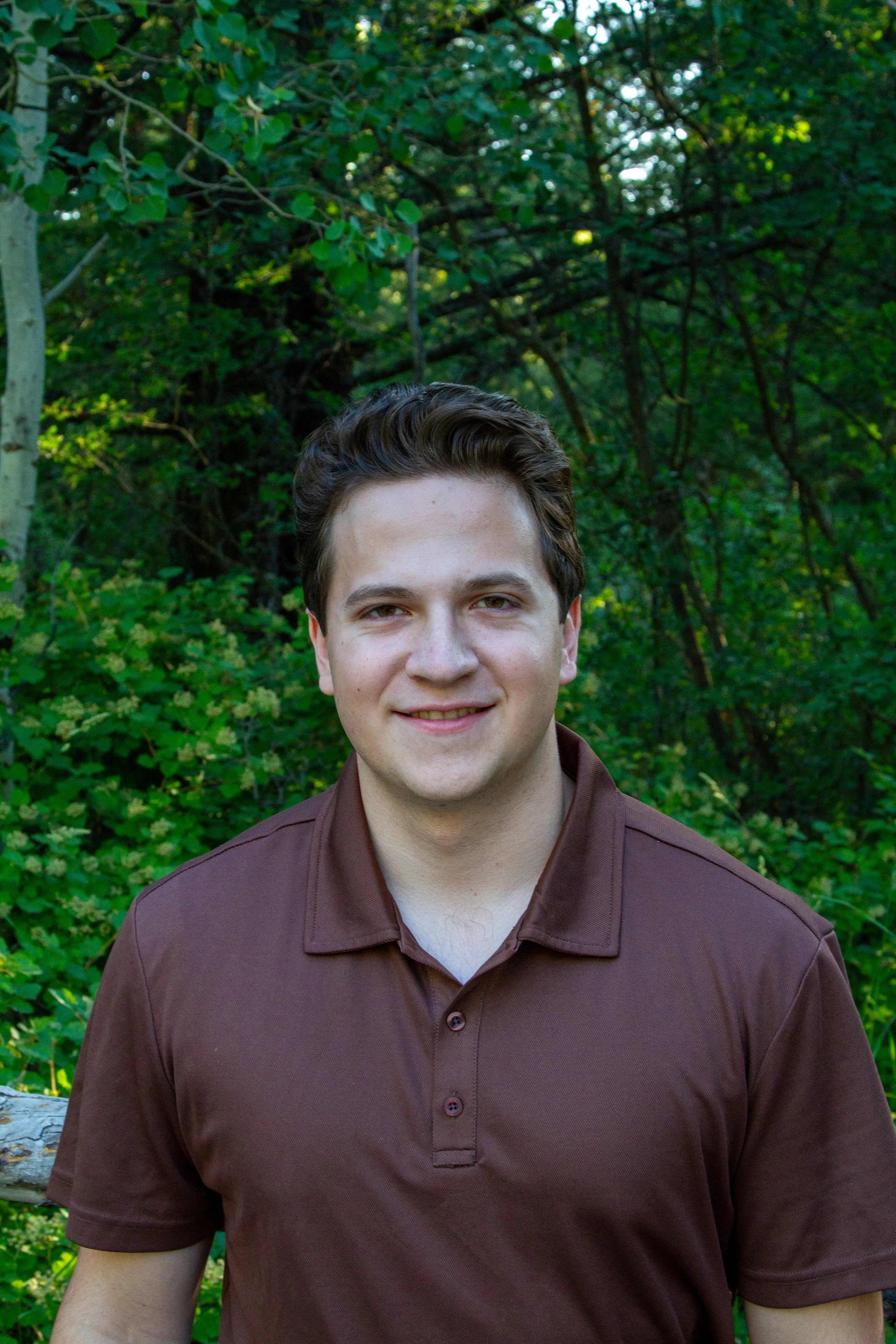 A young man with brown hair and light skin smiling outdoors in front of green trees and foliage, wearing a brown collared shirt.