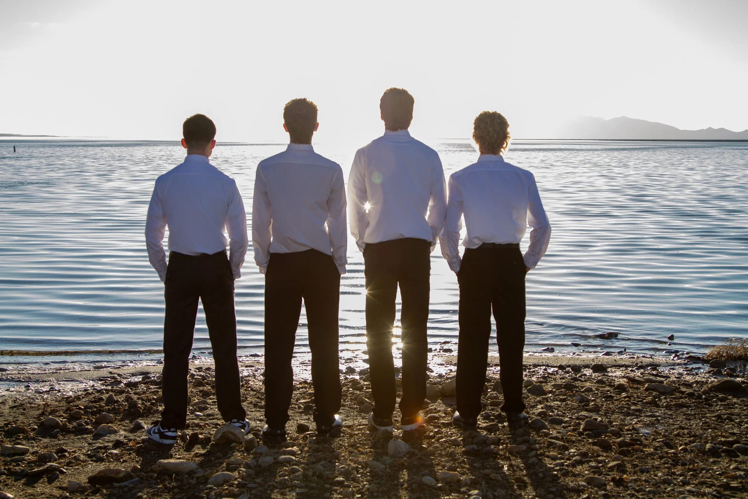 Four young men are standing on a rocky beach facing the water, wearing white shirts and dark pants, with a sunset or sunrise creating a silhouette effect.