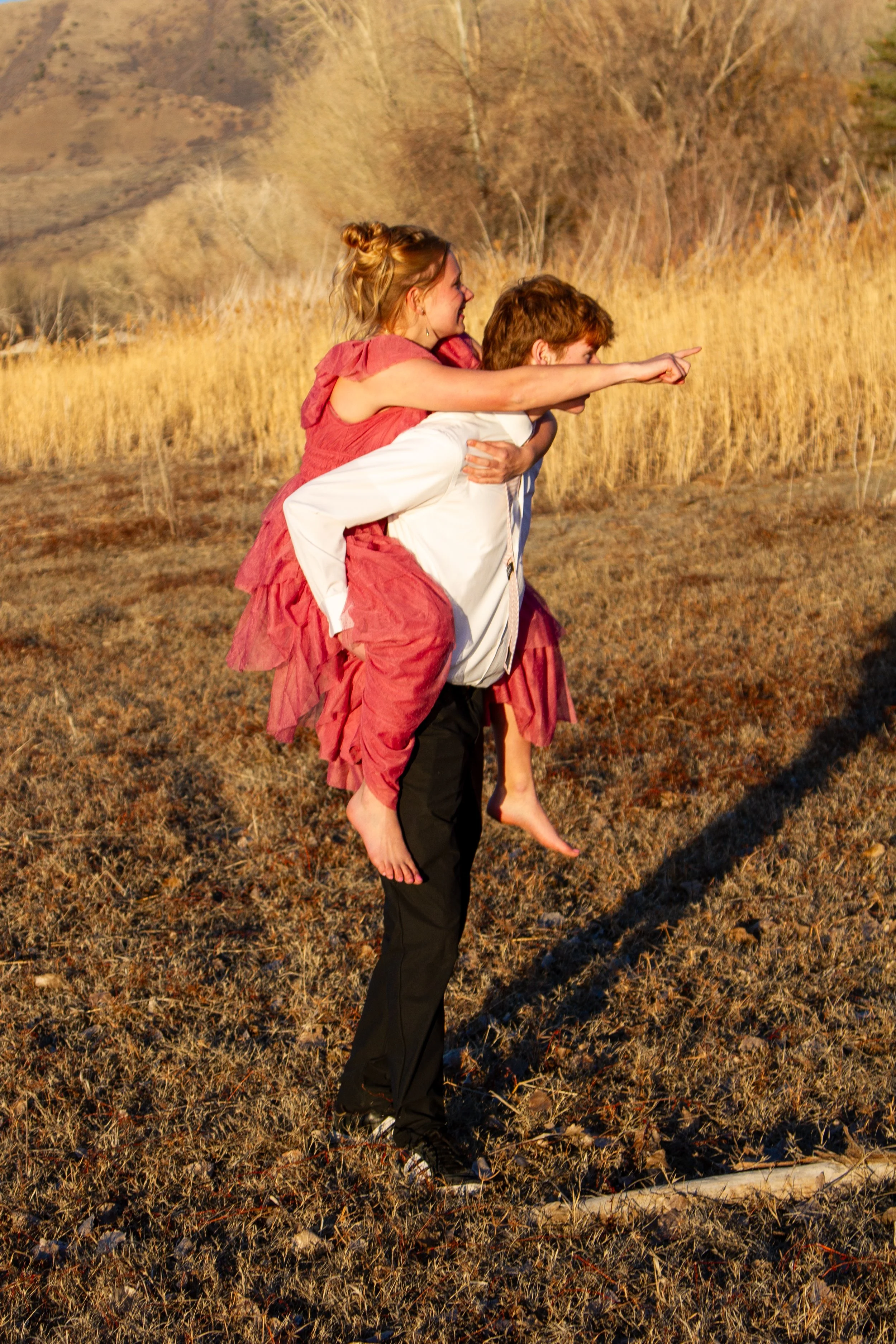 A man is standing outdoors in a dry, grassy field, carrying a young girl on his back who is pointing forward. The girl has her arms around the man's neck, and they are both smiling. The scene is lit by warm, golden sunlight, with trees and hills in t