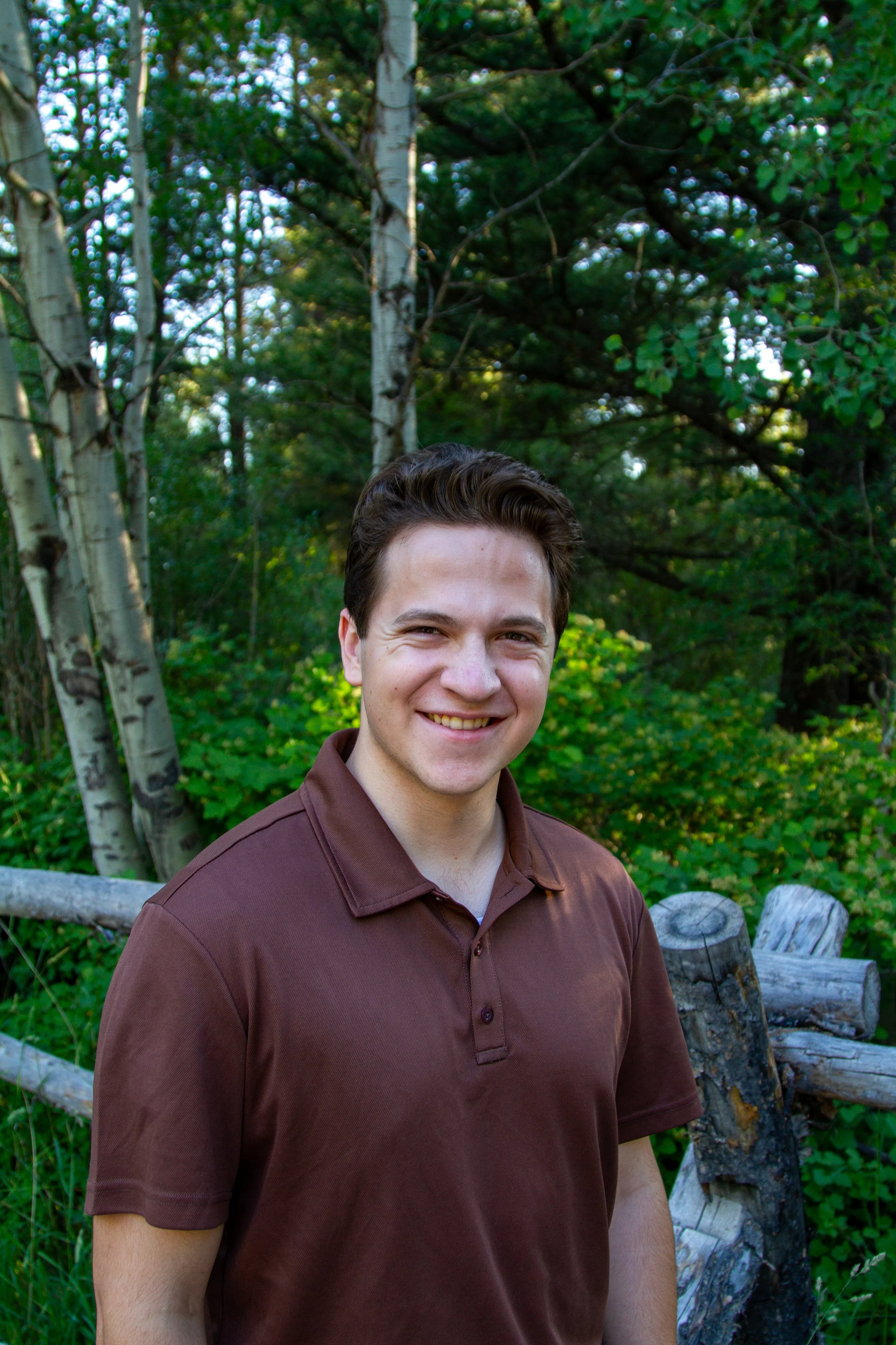 Young man with a broad smile, short brown hair, wearing a brown polo shirt, standing outdoors in front of green trees and logs.