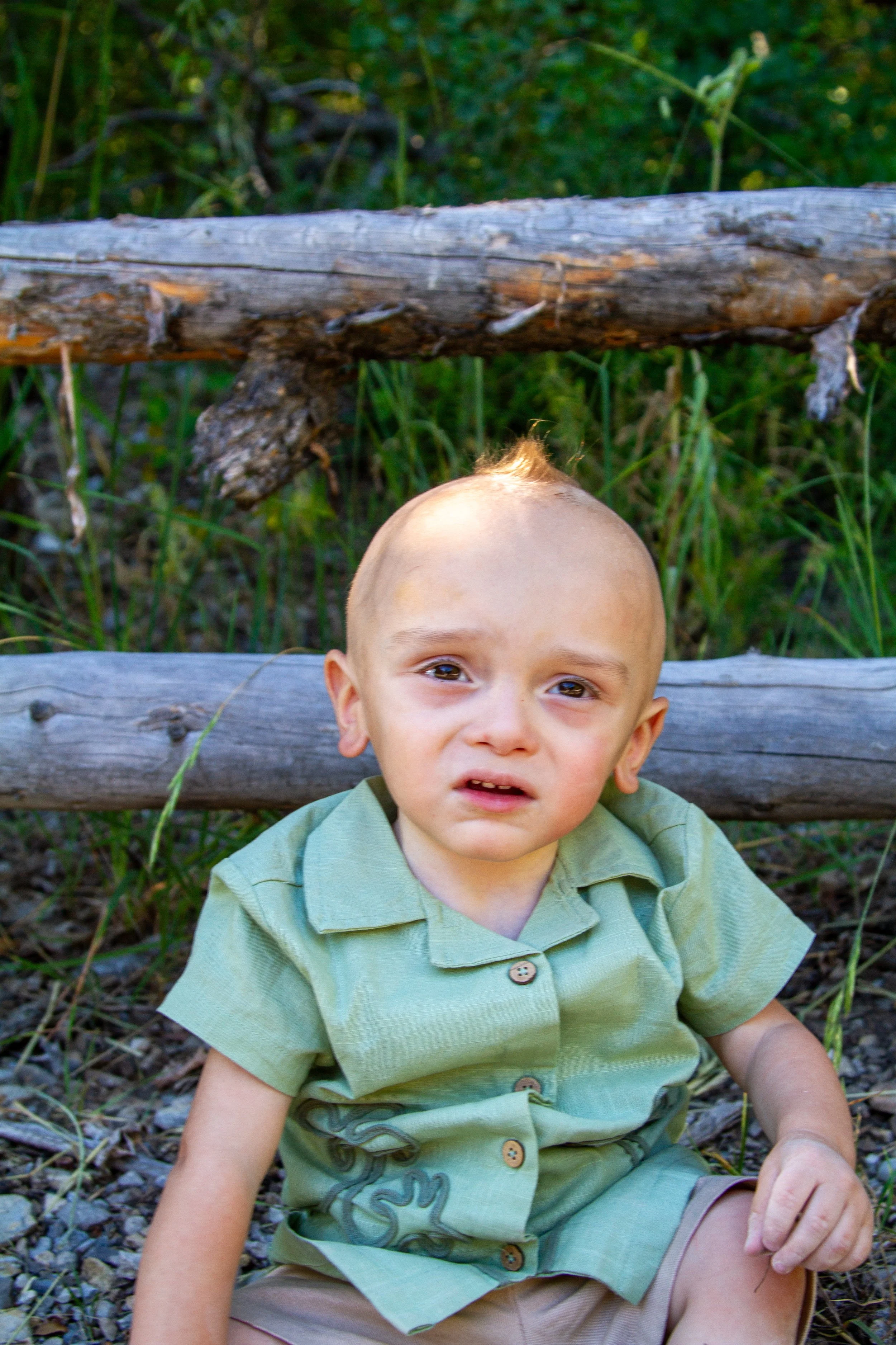 A young boy with a mohawk hairstyle sitting outdoors against a wooden fence with green grass and plants in the background.