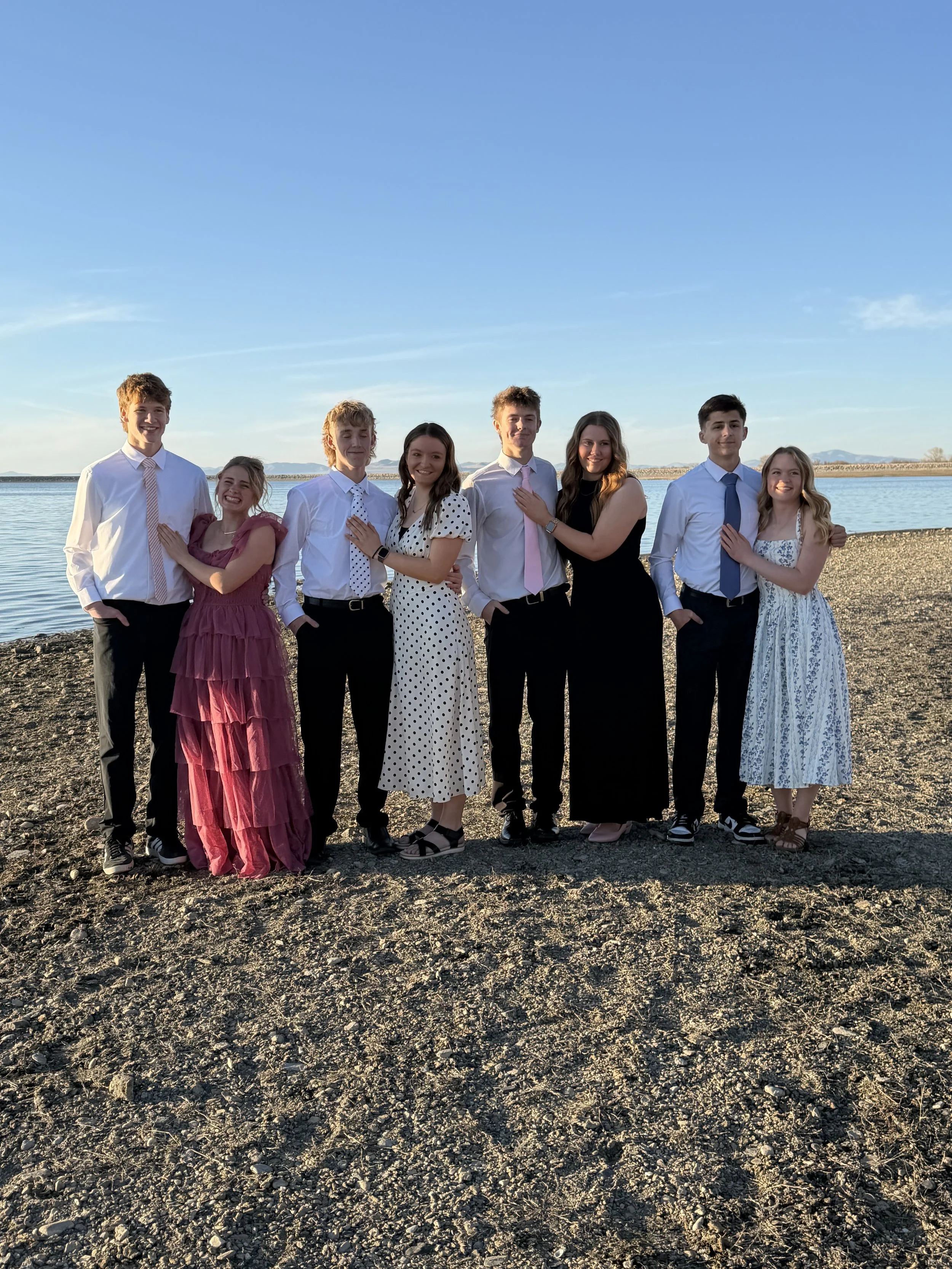 A group of eight young people dressed in formal and semi-formal attire standing on a gravelly shoreline by the water during sunset or late afternoon.