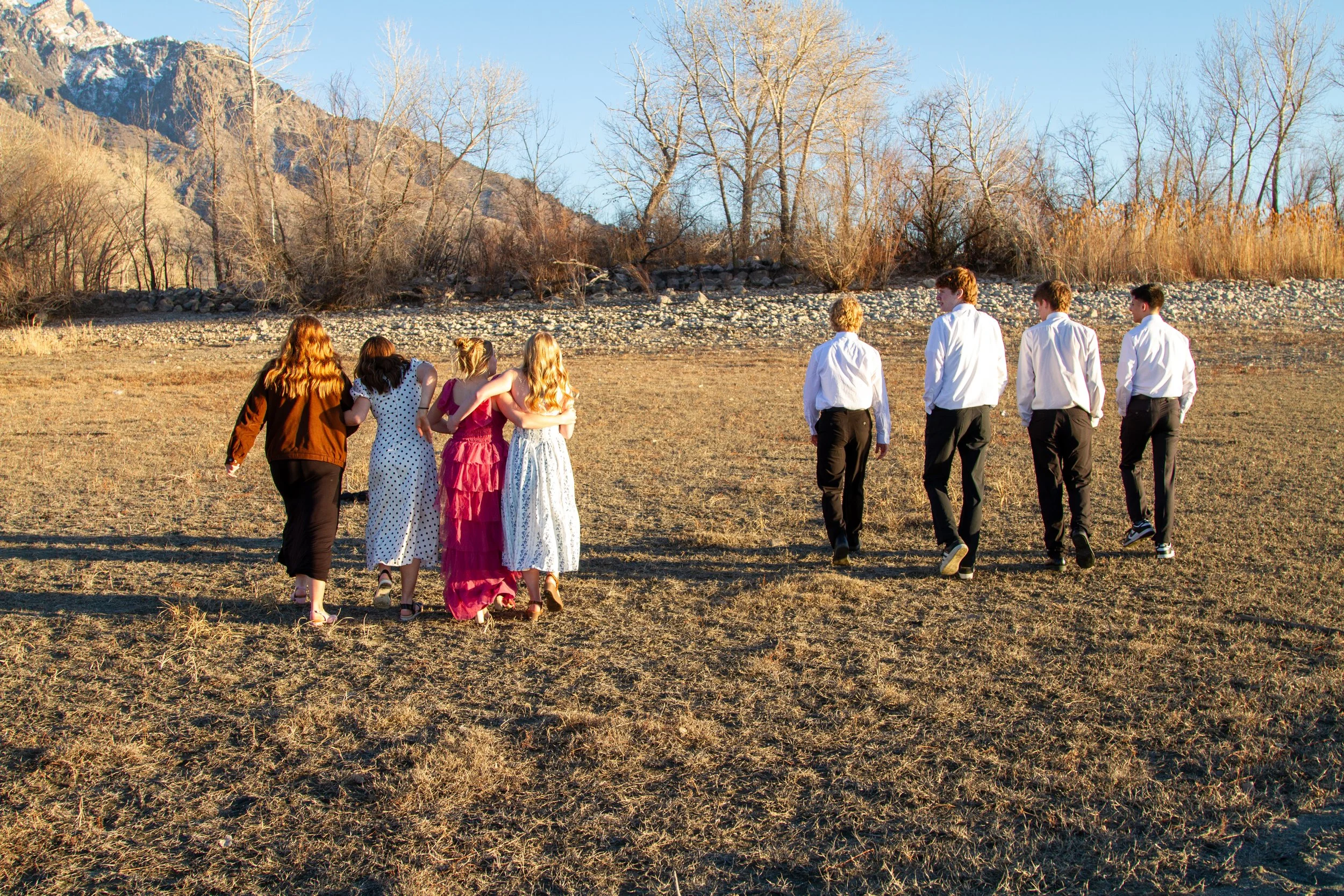 A group of seven people, three women and four men, walking away on a dry grassy field with mountains and leafless trees in the background during late afternoon or early evening.