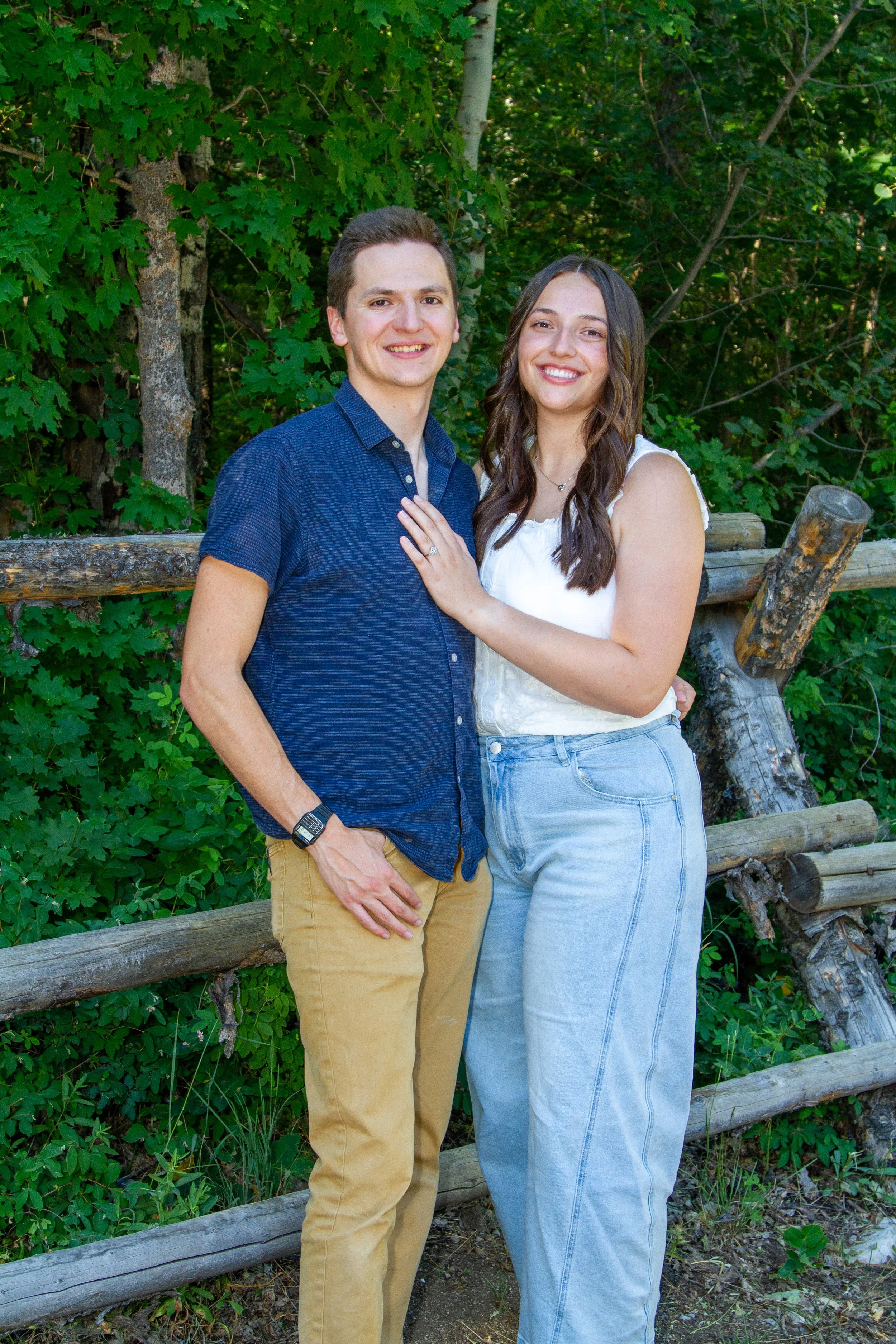 A young man and woman standing outdoors in front of a green leafy background. The man is wearing a blue button-up shirt and tan pants, and the woman is wearing a white sleeveless top and light blue jeans. They are smiling and the woman has her hand o