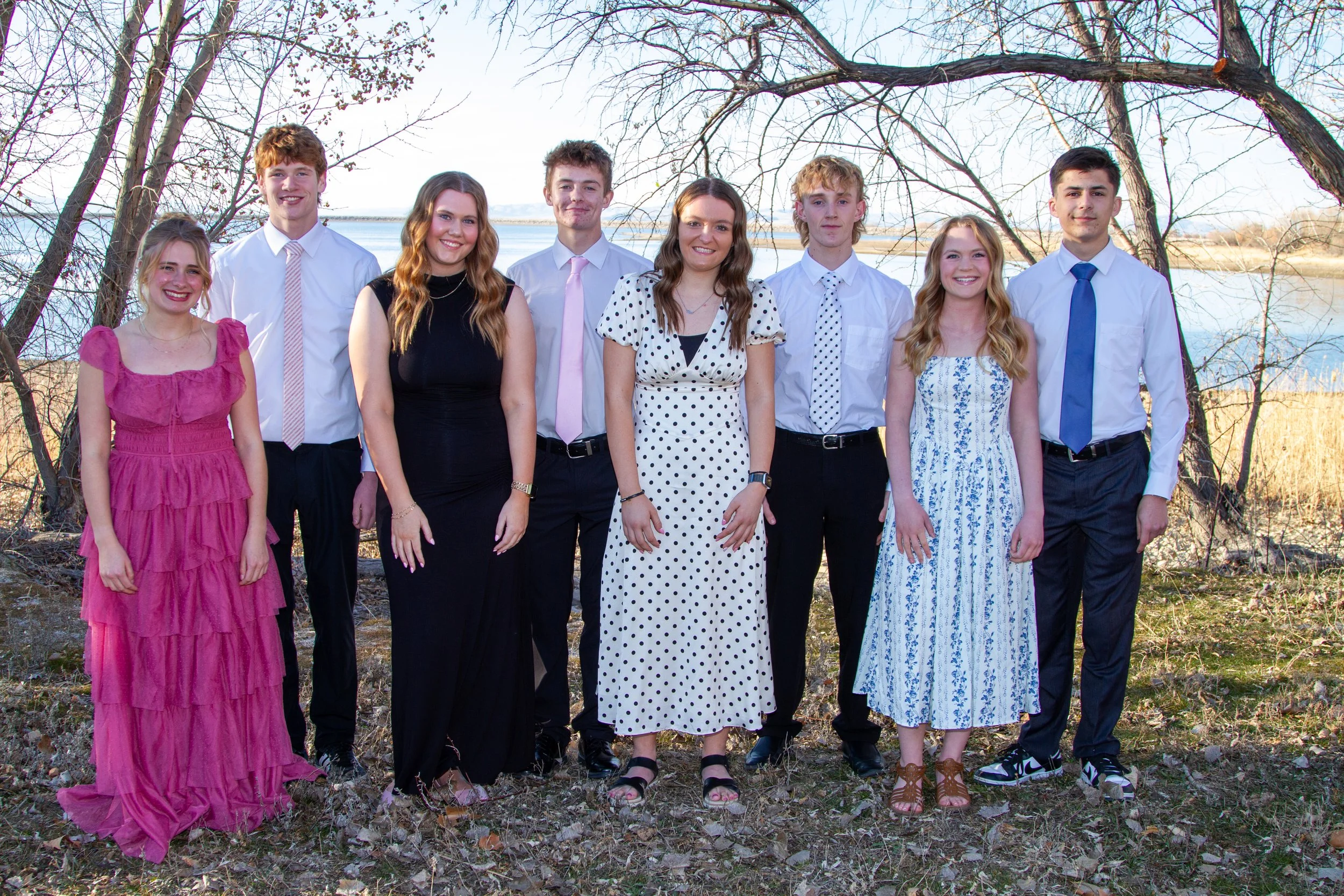 Group of nine young people standing outdoors near a lake, dressed in semi-formal attire, with trees in the background.