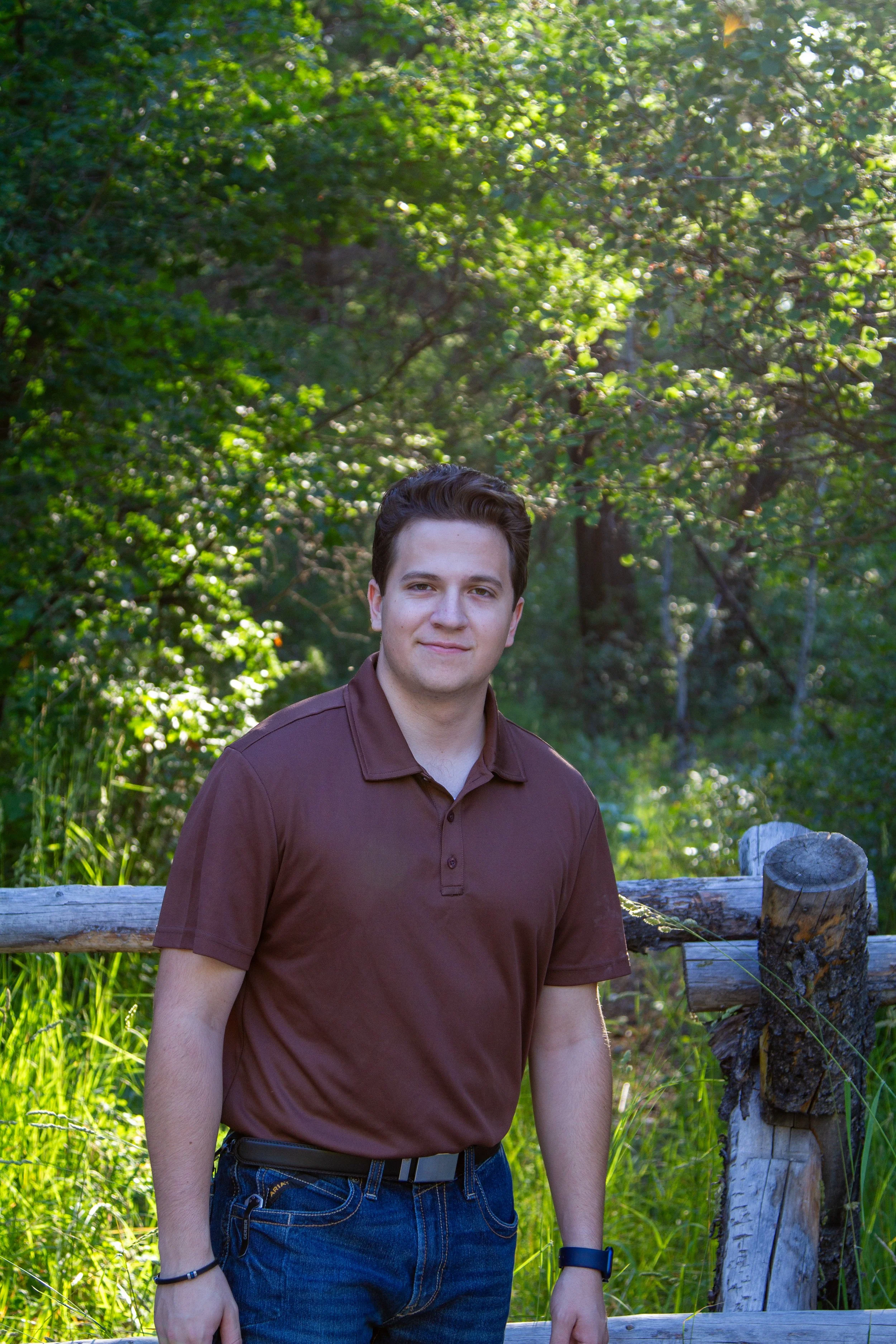 A young man standing outdoors in a park or forest, wearing a brown polo shirt, blue jeans, and a black wristwatch, with green trees and grass in the background.